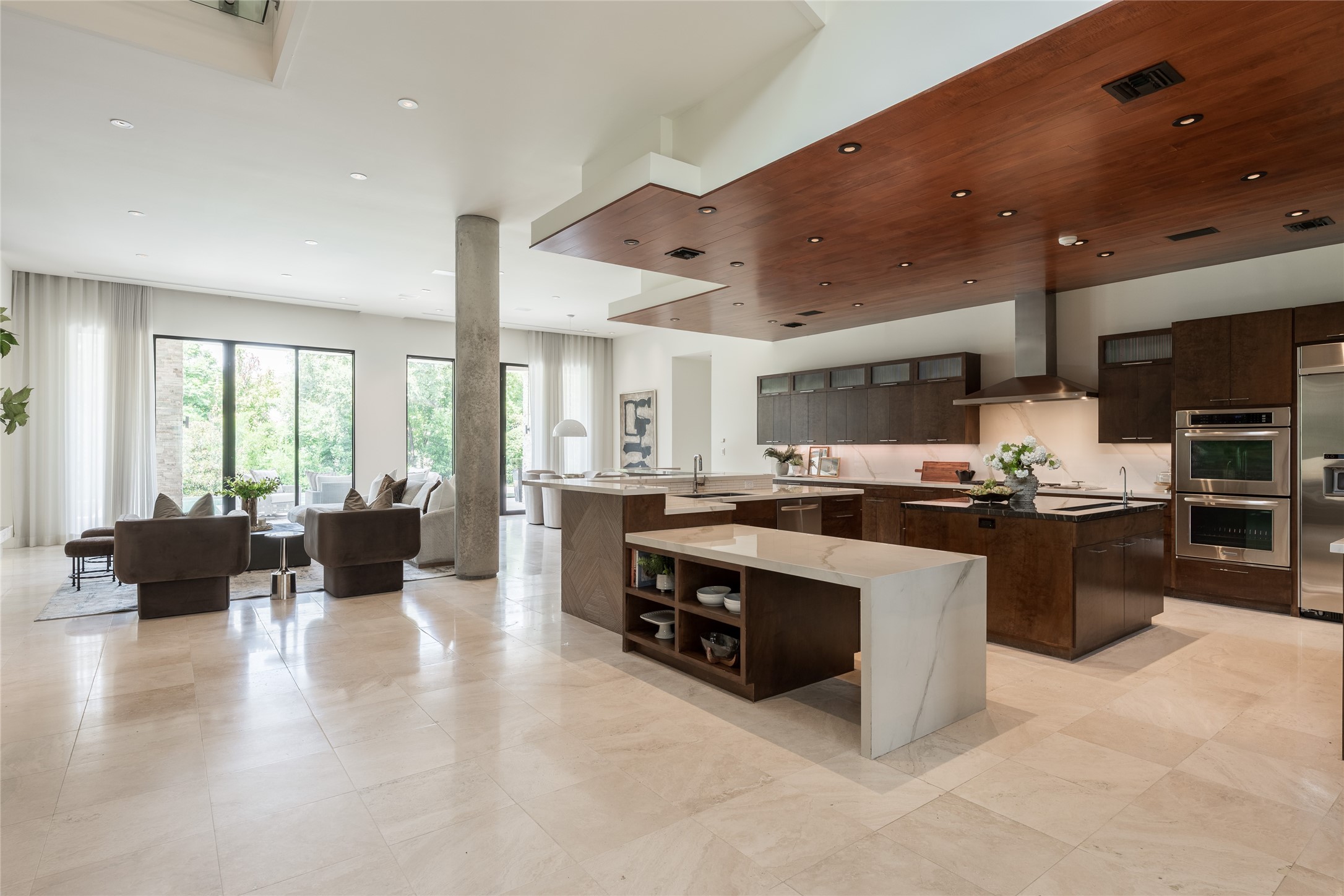 A suspended ceiling creates an architectural canopy over the kitchen, visually defining the space without separating it from the surrounding living areas. Custom cabinetry continues the material palette below, giving the kitchen a warmth and richness that contrasts beautifully against the homes lighter finishes