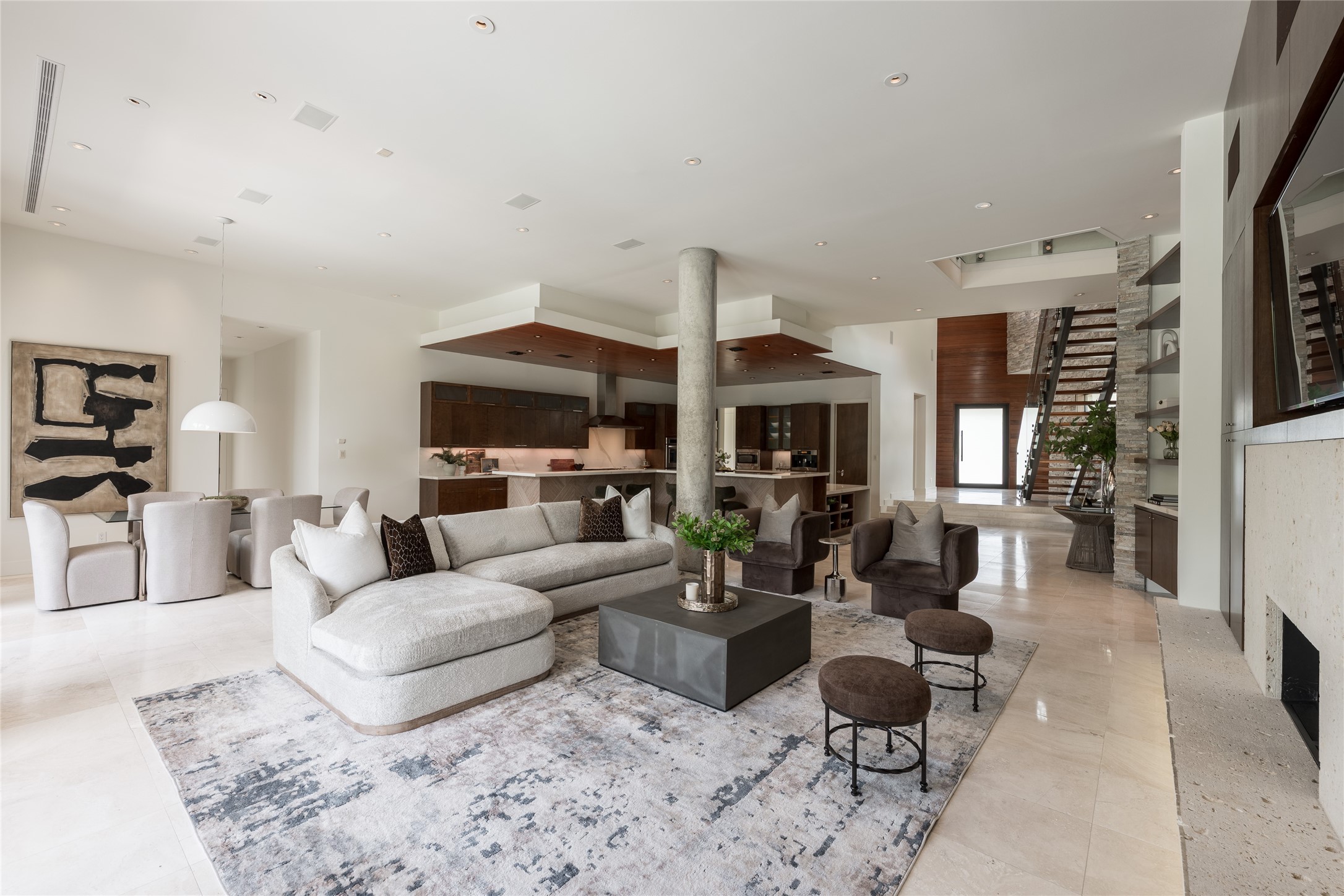 The main living room opens with impressive scale, where soaring ceilings and walls of glass behind fill the space with natural light. A prominent structural concrete column becomes an architectural feature rather than something concealed, adding a distinctive contrast to the homes warmer materials while helping define the transition between the living and kitchen spaces.