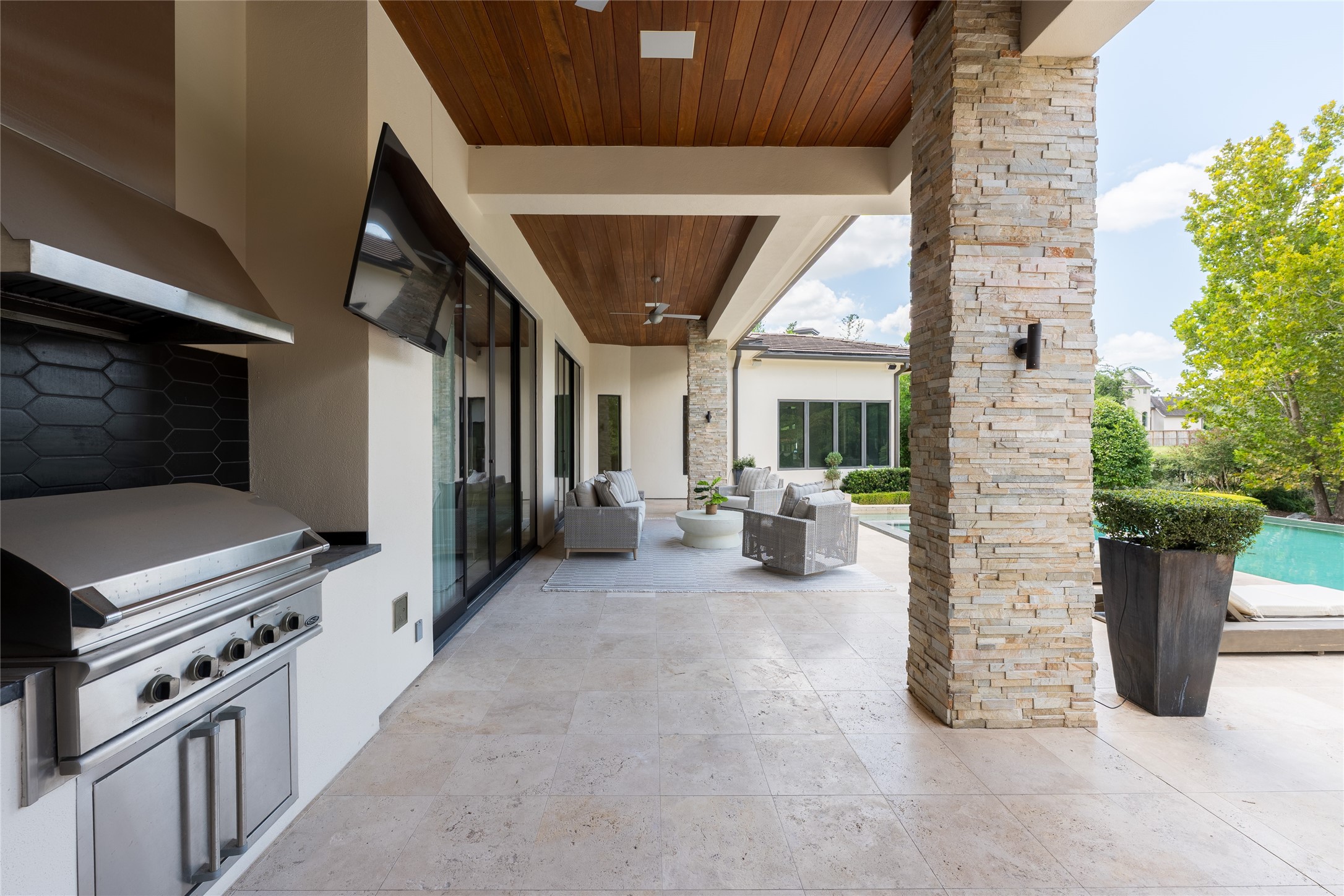The covered outdoor living area features a full summer kitchen beneath a mahogany tongue-and-groove ceiling, while the homes travertine flooring continues outdoors for a seamless transition in materials. Positioned alongside the pool, the space is designed for year-round cooking, dining and entertaining.