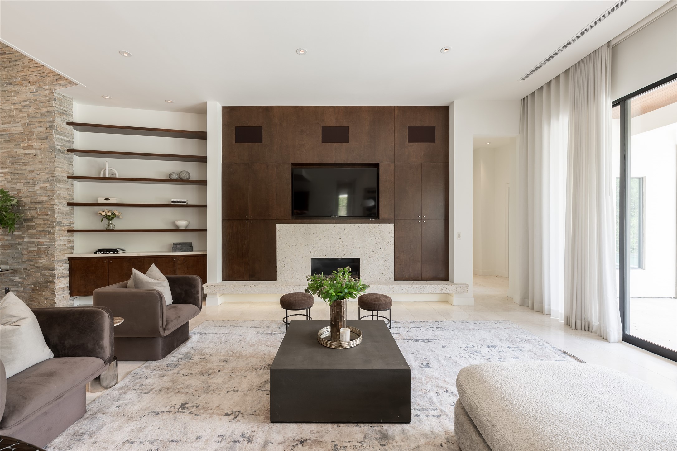 The living room is anchored by a natural travertine fireplace, framed by floating shelves and custom walnut cabinetry that bring warmth and symmetry to the space. Hidden speakers positioned above the cabinetry integrate the surround-sound system discreetly into the architecture, keeping the technology heard but virtually unseen.