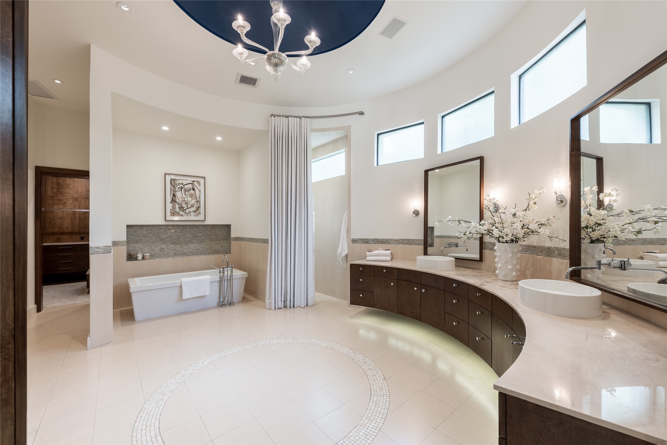 The primary ensuite showcases custom floating curved cabinetry that follows the architecture of the room, centered by a freestanding soaking tub above a mosaic-tiled cove as an oversized walk in shower with dual showers is nestled discreetly.
