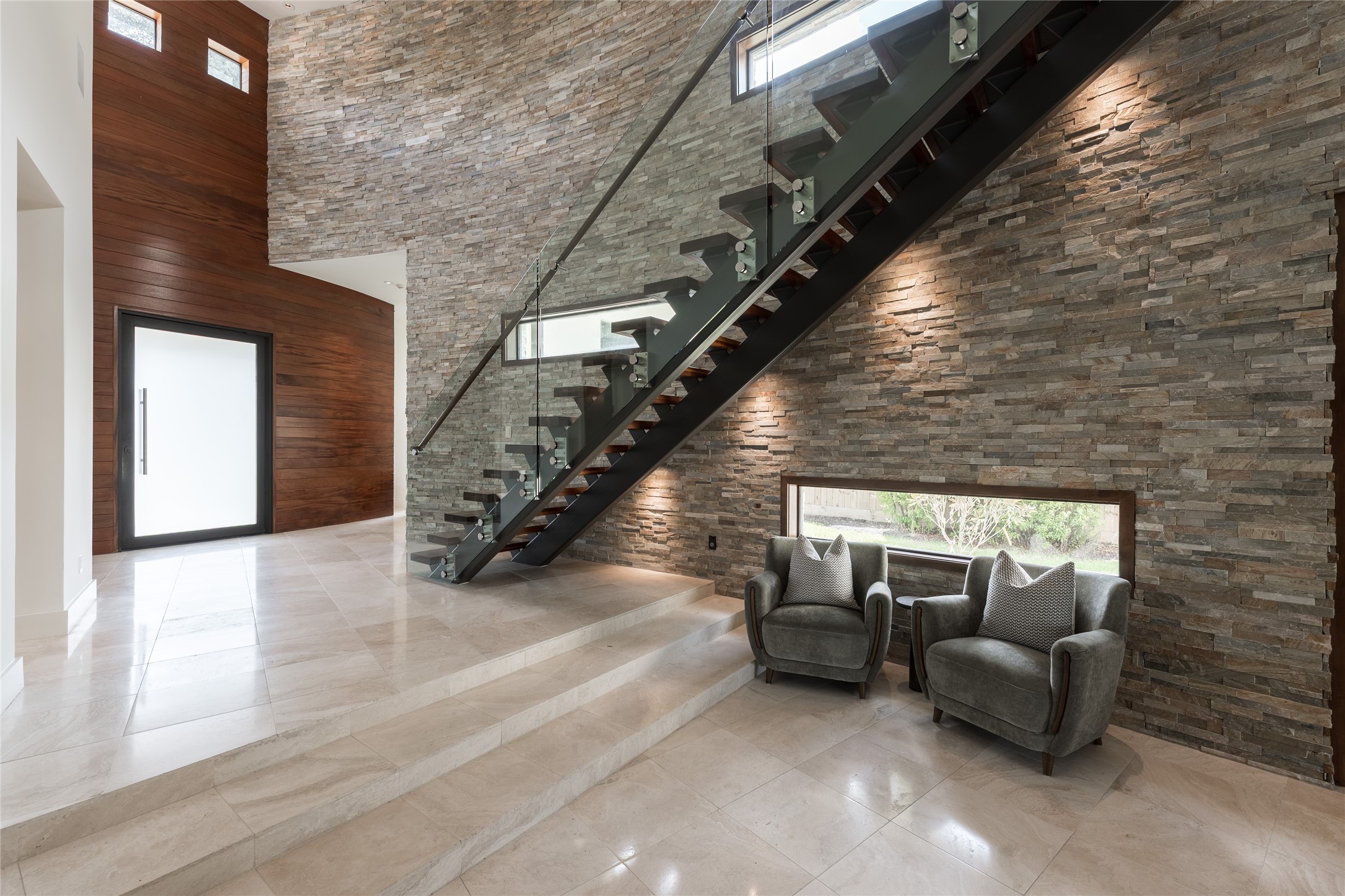 The foyer creates a dramatic sense of arrival through a mahogany-framed 60' pivot door, polished travertine floors and a sculptural curved stacked-stone wall. A subtle step-down transitions from the entry into the main residence, revealing the home gradually rather than all at once.