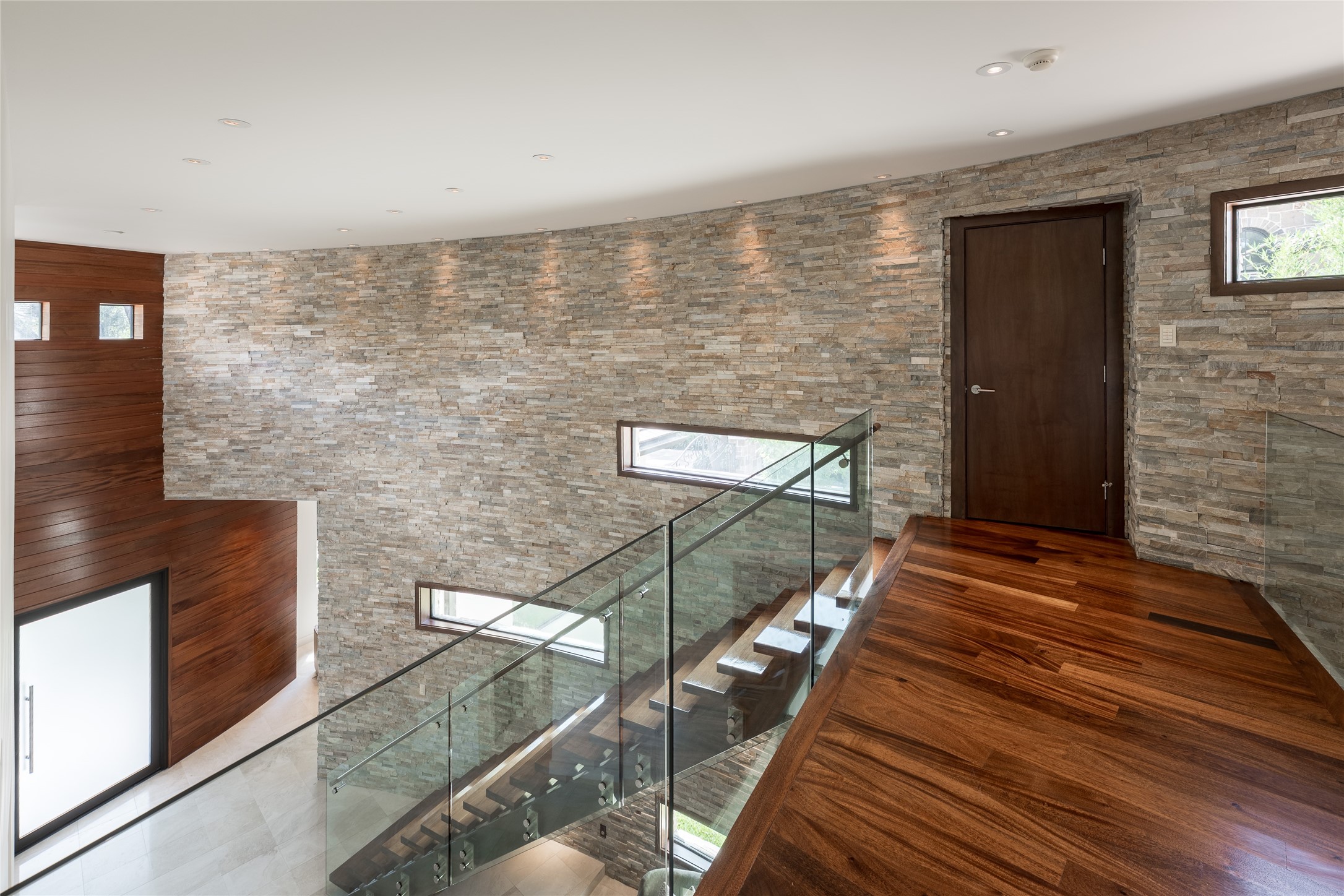 The upstairs landing continues the homes curved stacked-stone wall and walnut flooring, overlooking the floating staircase below. A dedicated storage closet is integrated along the landing, adding practical storage without interrupting the open architectural design.
