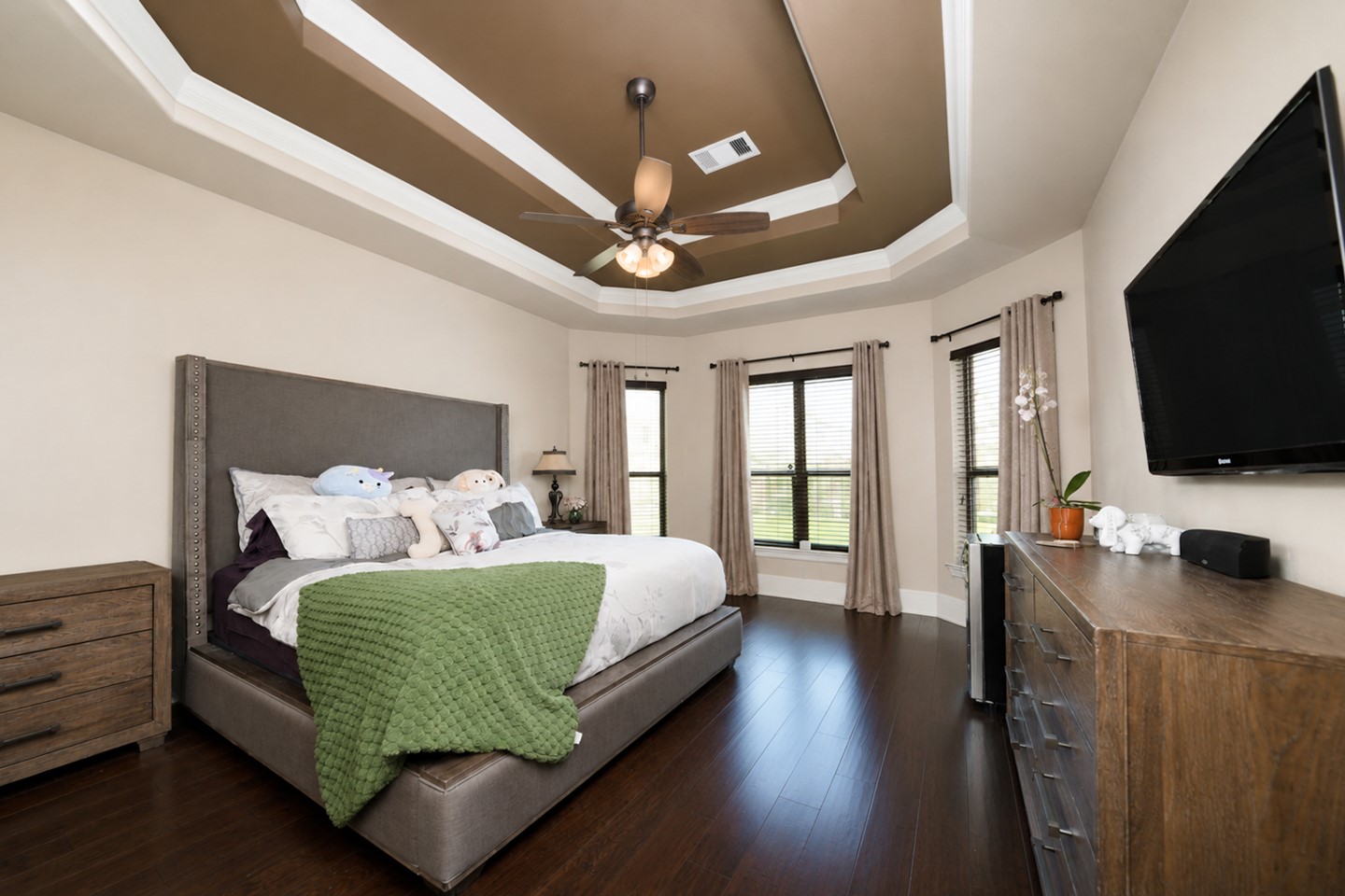 Spacious primary bedroom with elegant tray ceiling and ceiling fan, featuring hardwood floors and ample natural light from large windows.