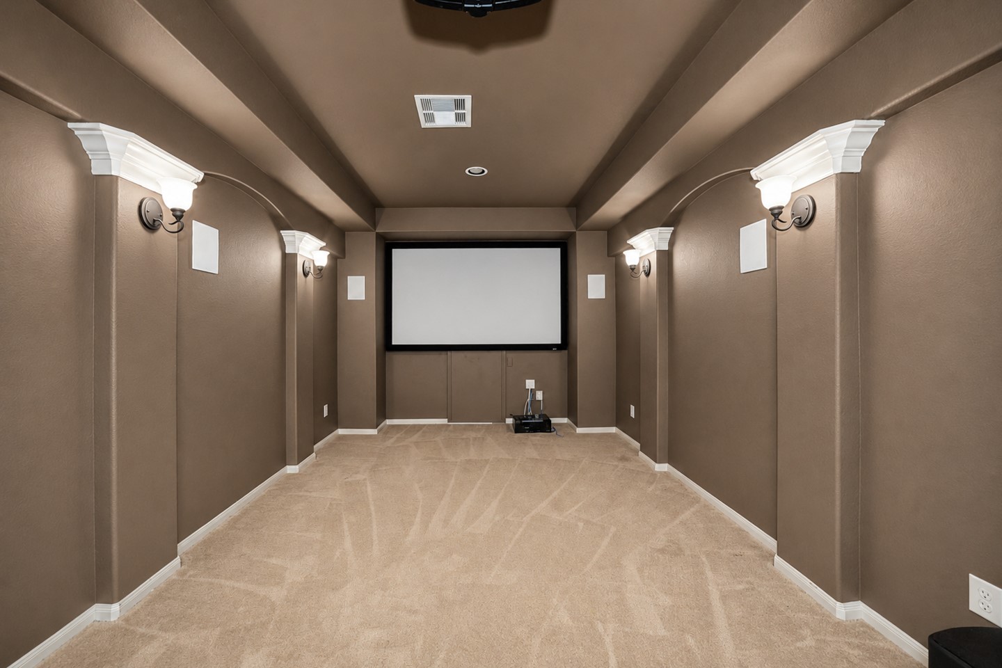 This photo shows a cozy home theater room with a large screen, carpeted flooring, and elegant wall sconces for ambient lighting. The neutral tones create a warm and inviting atmosphere, perfect for movie nights.