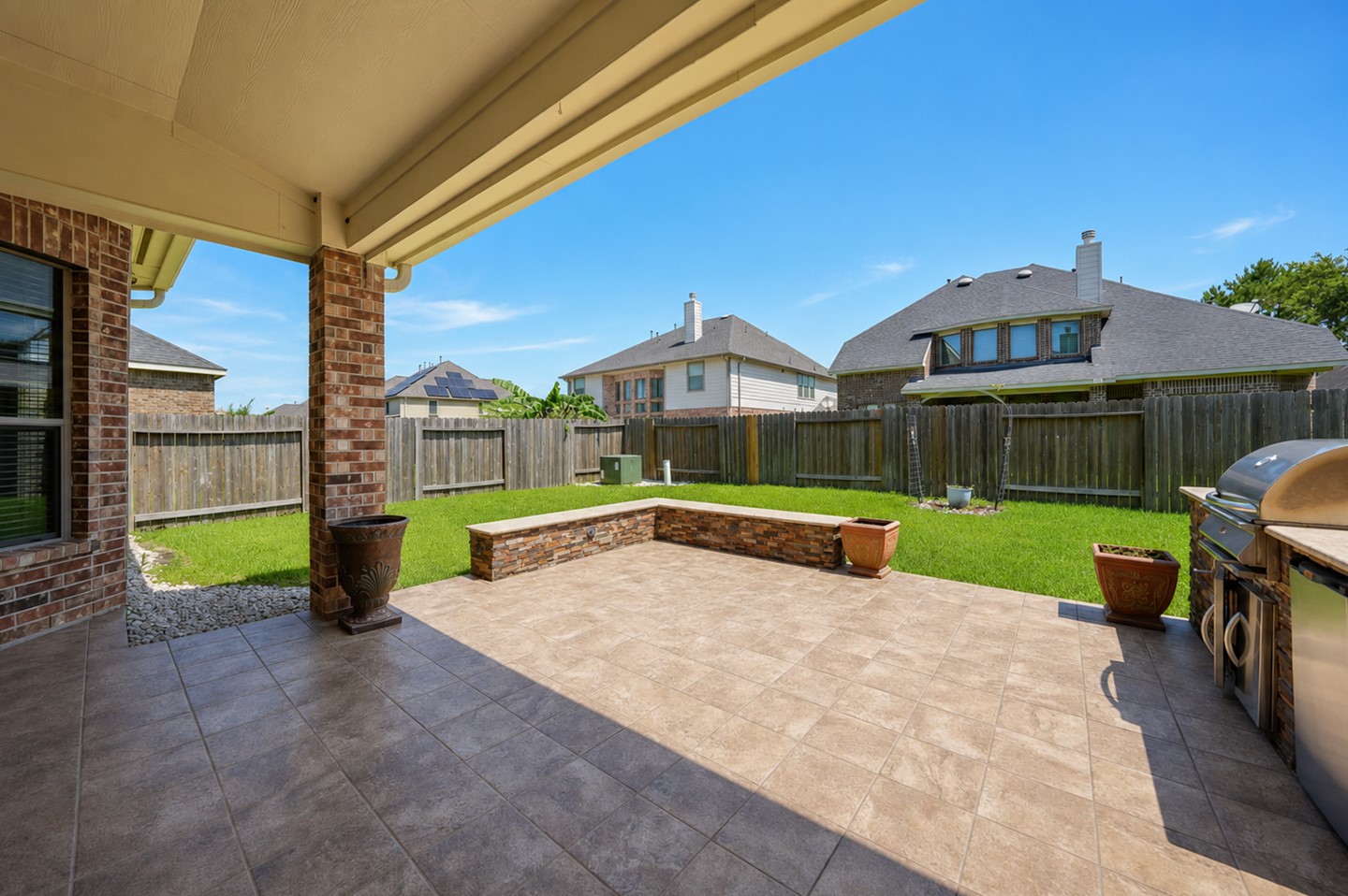 This photo showcases a spacious, covered patio with tiled flooring, perfect for outdoor entertainment. It includes a built-in grill and overlooks a well-maintained, fenced backyard with a small stone bench, providing privacy and a great space for relaxation.