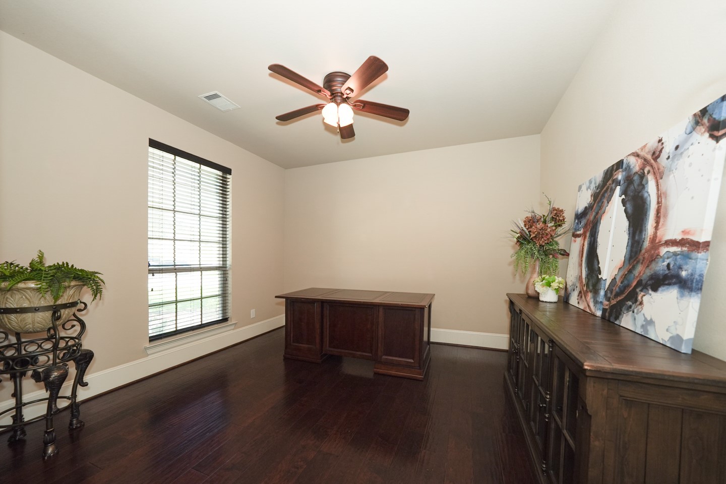 This photo features a bright, spacious office or study space with dark wood flooring and neutral walls. It includes a ceiling fan with a large window.