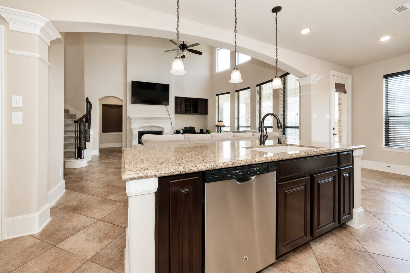This modern open-concept living space features a spacious kitchen with granite countertops, pendant lighting, and stainless-steel appliances. It flows into a bright living area with high ceilings, large windows, and a fireplace, creating a welcoming atmosphere.