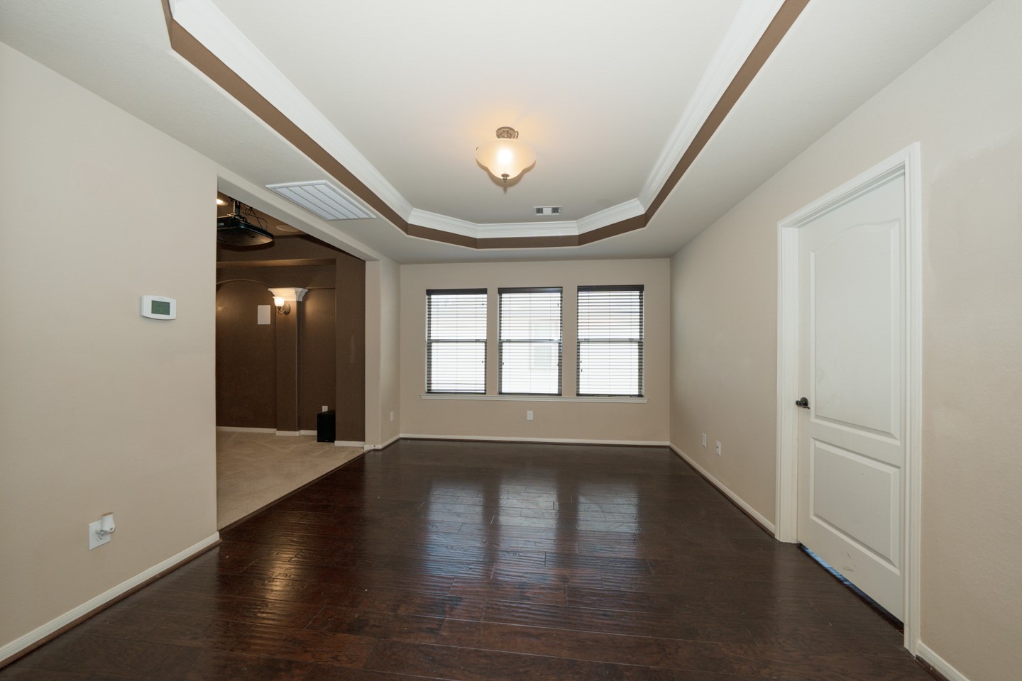 This flex room features dark hardwood floors, a tray ceiling and a large window for natural light. It offers a spacious, open feel with an adjacent area visible through a wide doorway.