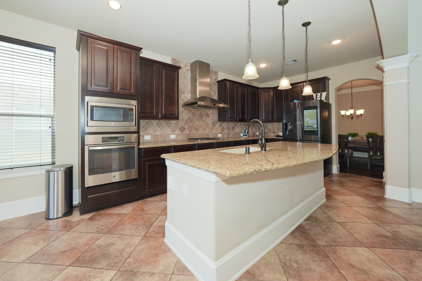 This modern kitchen features dark wood cabinets, stainless steel appliances, and a large granite-topped island with pendant lighting. The open layout flows into a dining area, highlighted by tiled floors and ample natural light.