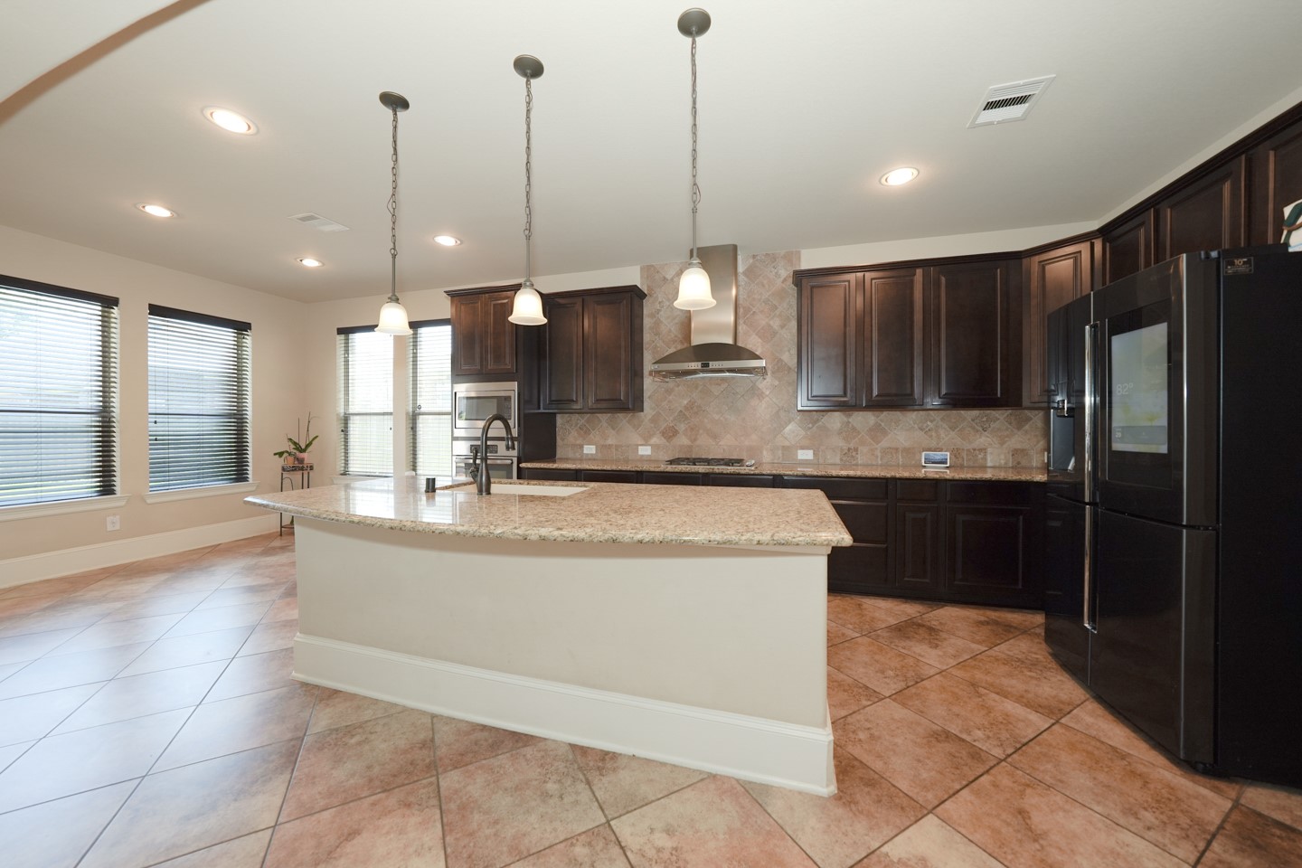 This spacious kitchen features dark wood cabinetry, a large granite island and pendant lighting. The open layout is accented by tiled flooring and ample natural light from multiple windows.