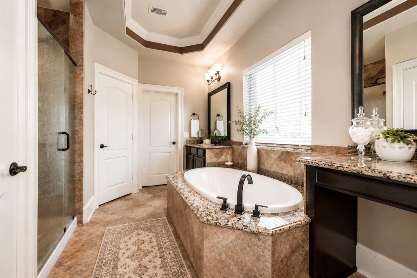 This elegant primary bathroom features a central soaking tub with granite surround, a spacious glass-enclosed shower, dual vanities with large mirrors, and tasteful tile flooring. Natural light filters through a window with blinds, creating a bright and inviting space.