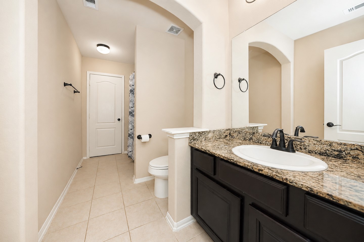 This bathroom features a spacious layout with a granite countertop, dark wood vanity, and a large mirror. It has a tiled floor and the Neutral tones create a warm and inviting atmosphere.