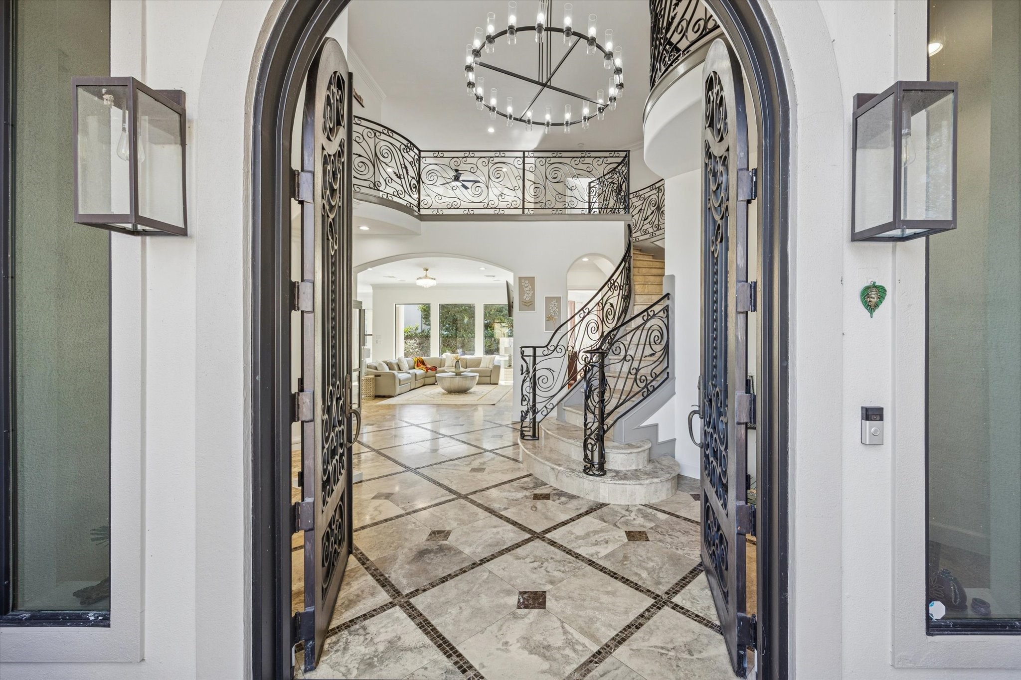 This elegant entryway features double iron doors opening to a grand foyer with a curved staircase and intricate wrought iron railings. The space is bright and open, with a large chandelier and travertine and marble inlay flooring, leading into a spacious living area with ample natural light.