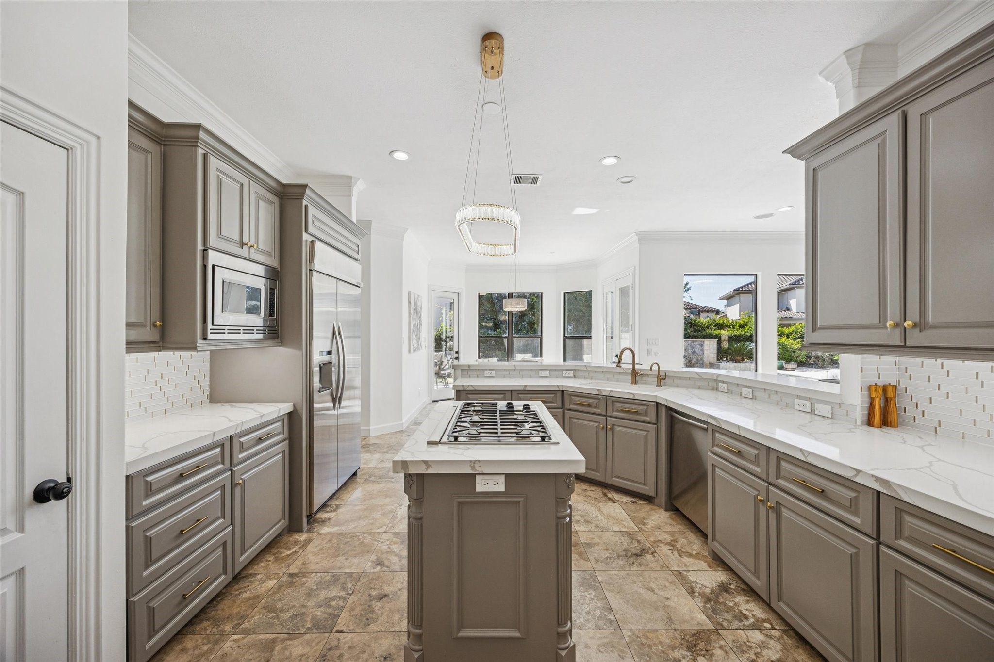 This spacious kitchen features elegant gray cabinetry, a large central island with a gas cooktop, and modern stainless steel appliances. The room is brightened by ample natural light from large windows, highlighting the sleek tile flooring and contemporary lighting fixture.