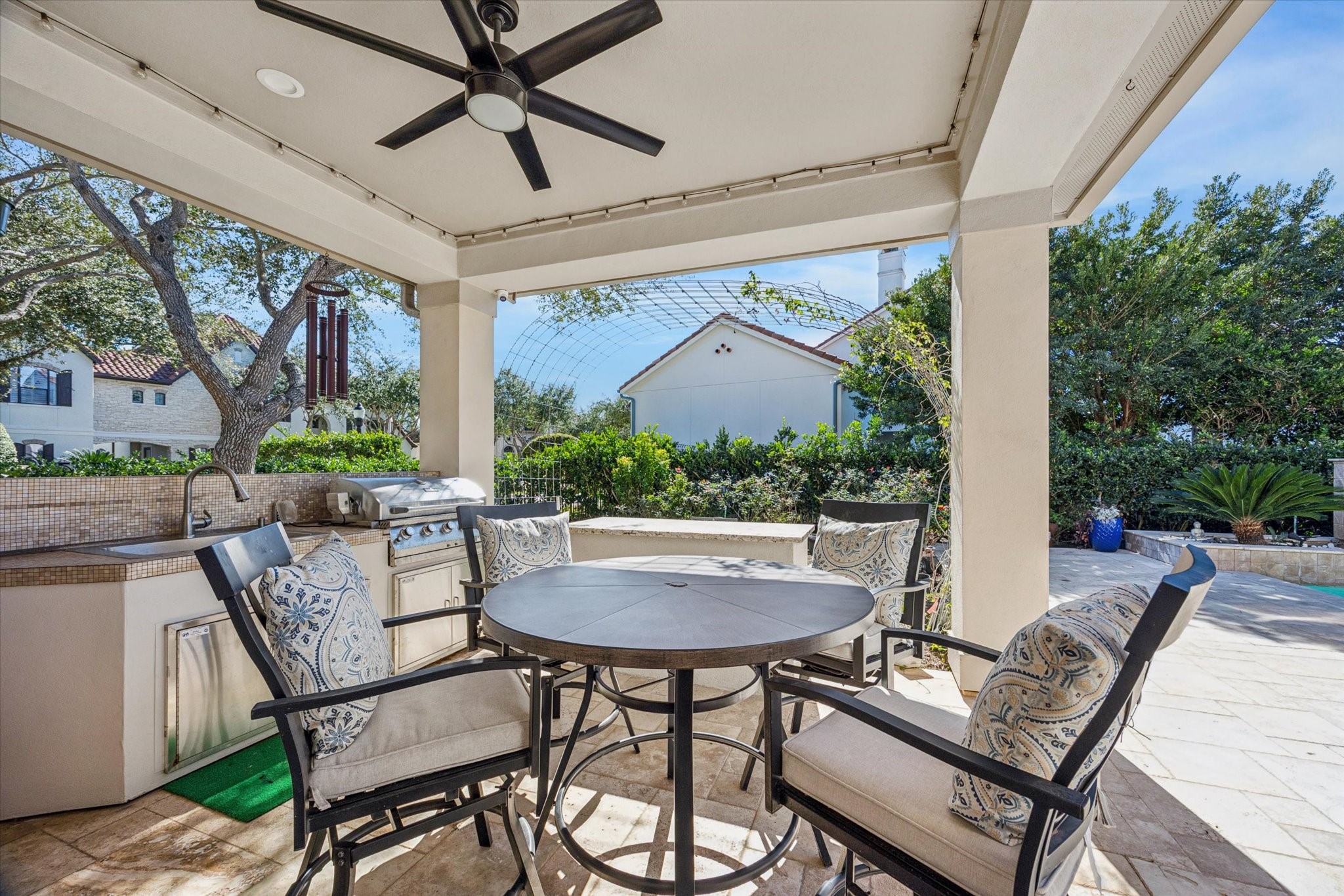 This photo showcases a stylish outdoor patio with a covered seating area, It features an outdoor kitchen with a built-in grill and sink, surrounded by lush greenery. The space offers a perfect setting for entertaining and relaxation.