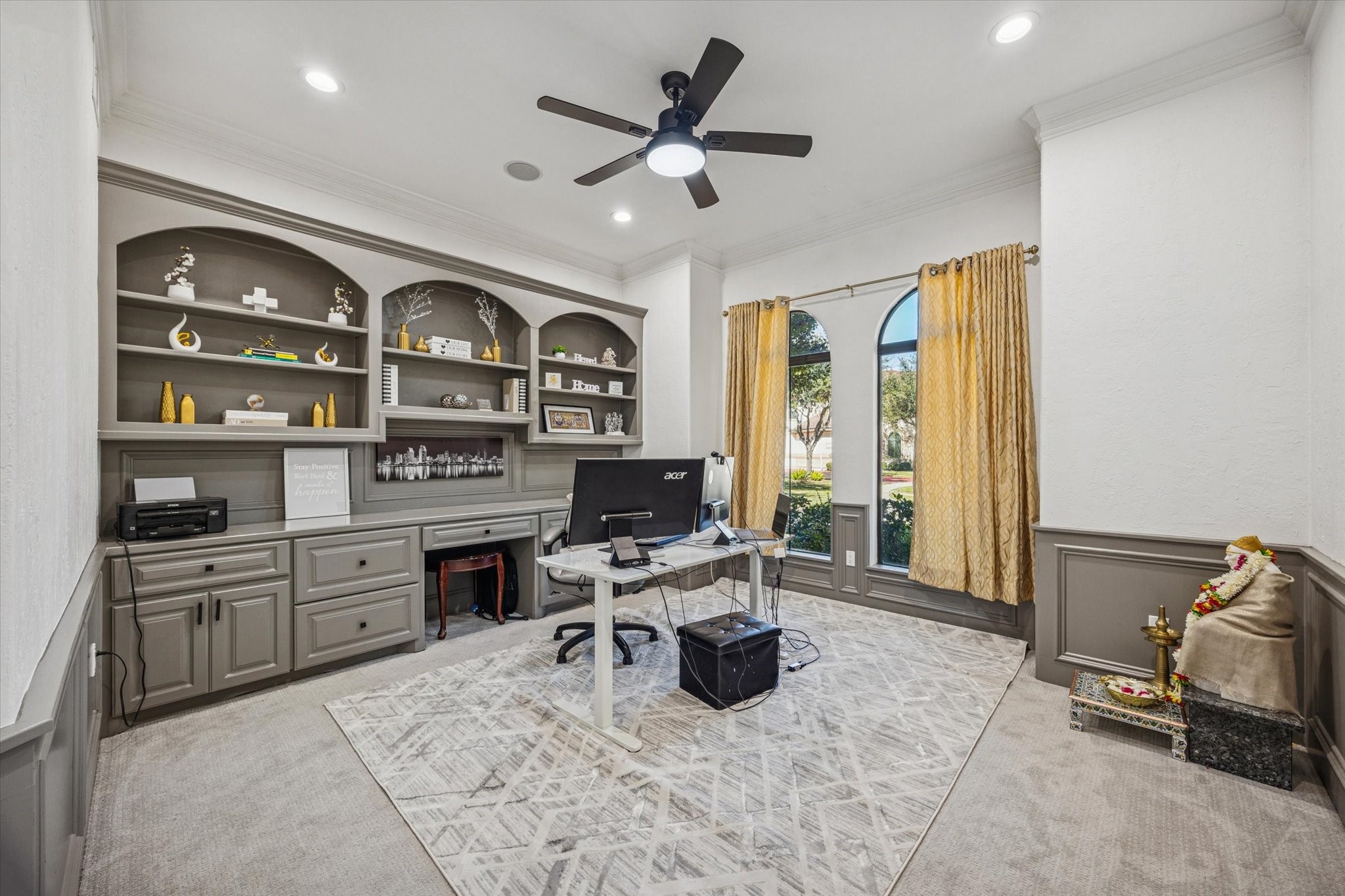 This photo showcases a bright home office with built-in shelves and cabinets, a ceiling fan, large windows with drapes, and neutral-colored walls and carpet. Ideal for a functional workspace.