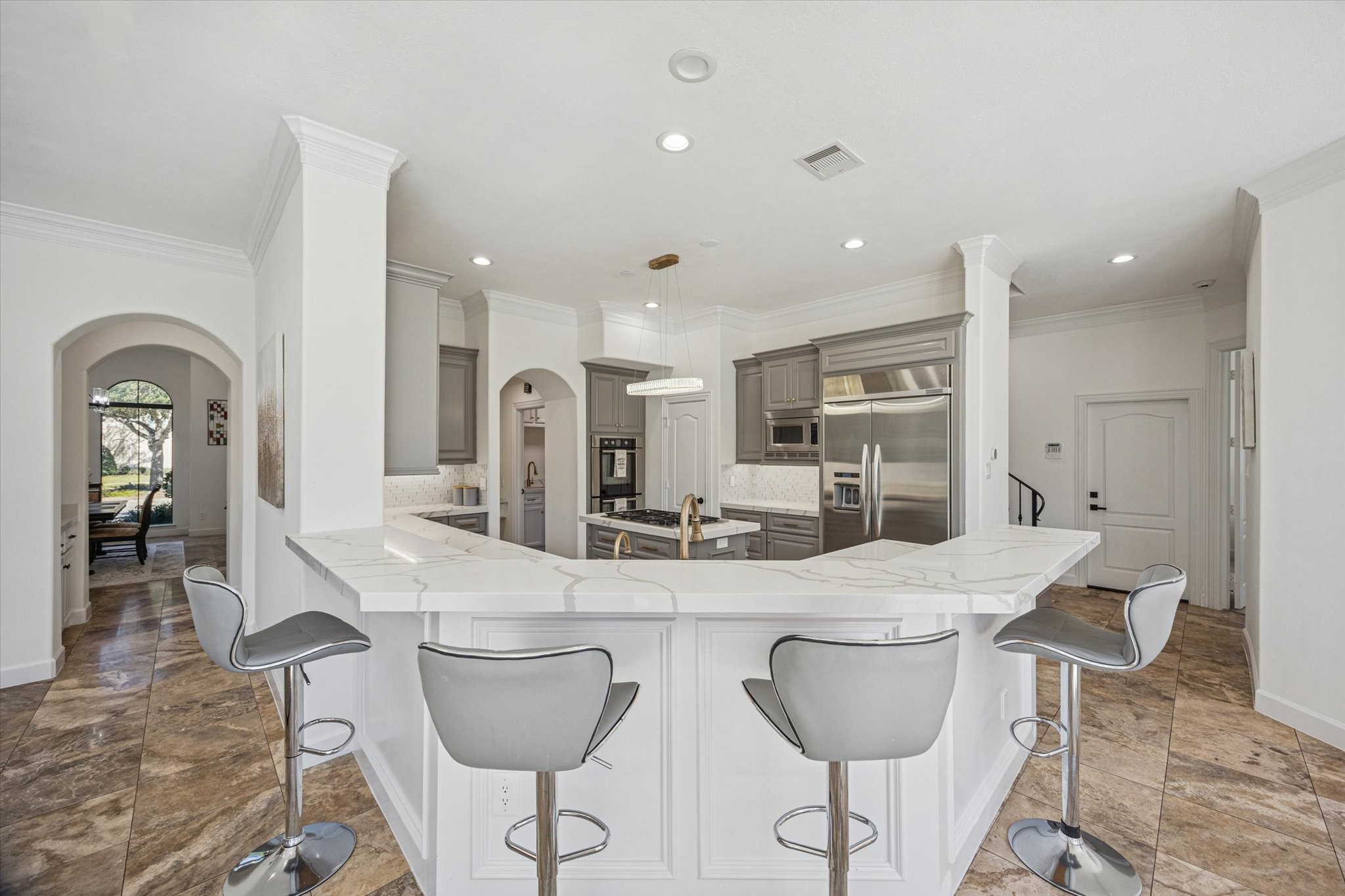 This modern kitchen features a spacious, open design with a large island and sleek bar seating. It includes stainless steel appliances, elegant cabinetry, and ample counter space, complemented by recessed lighting and tile flooring. An arched entryway leads to an adjacent room, enhancing the open flow.