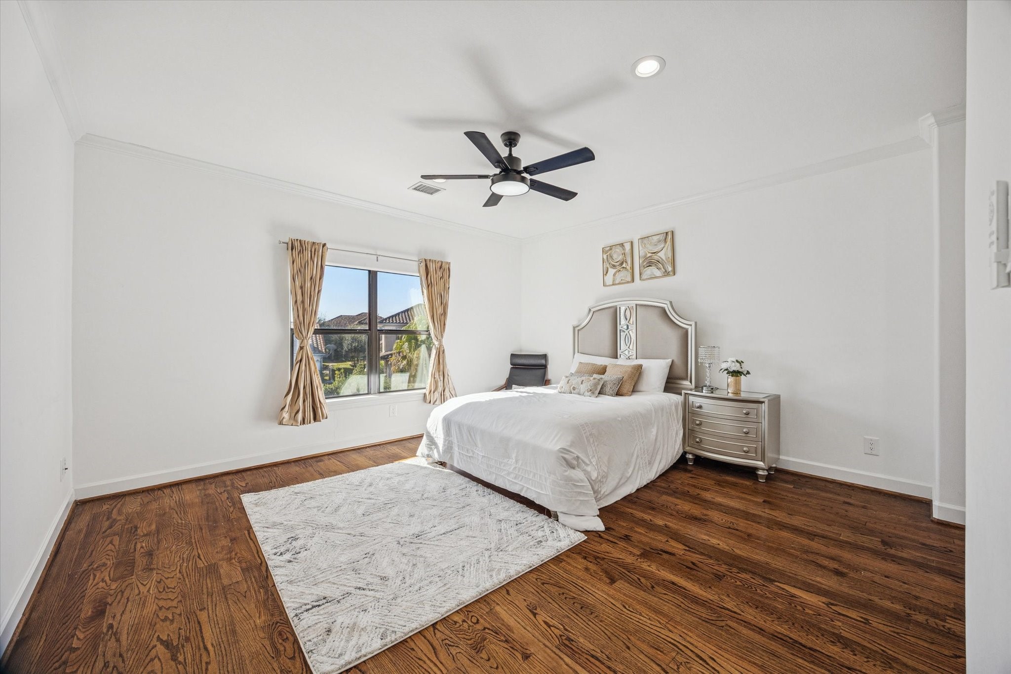 This is a bright, spacious upstairs bedroom featuring hardwood floors, a ceiling fan, and a large window with drapes.