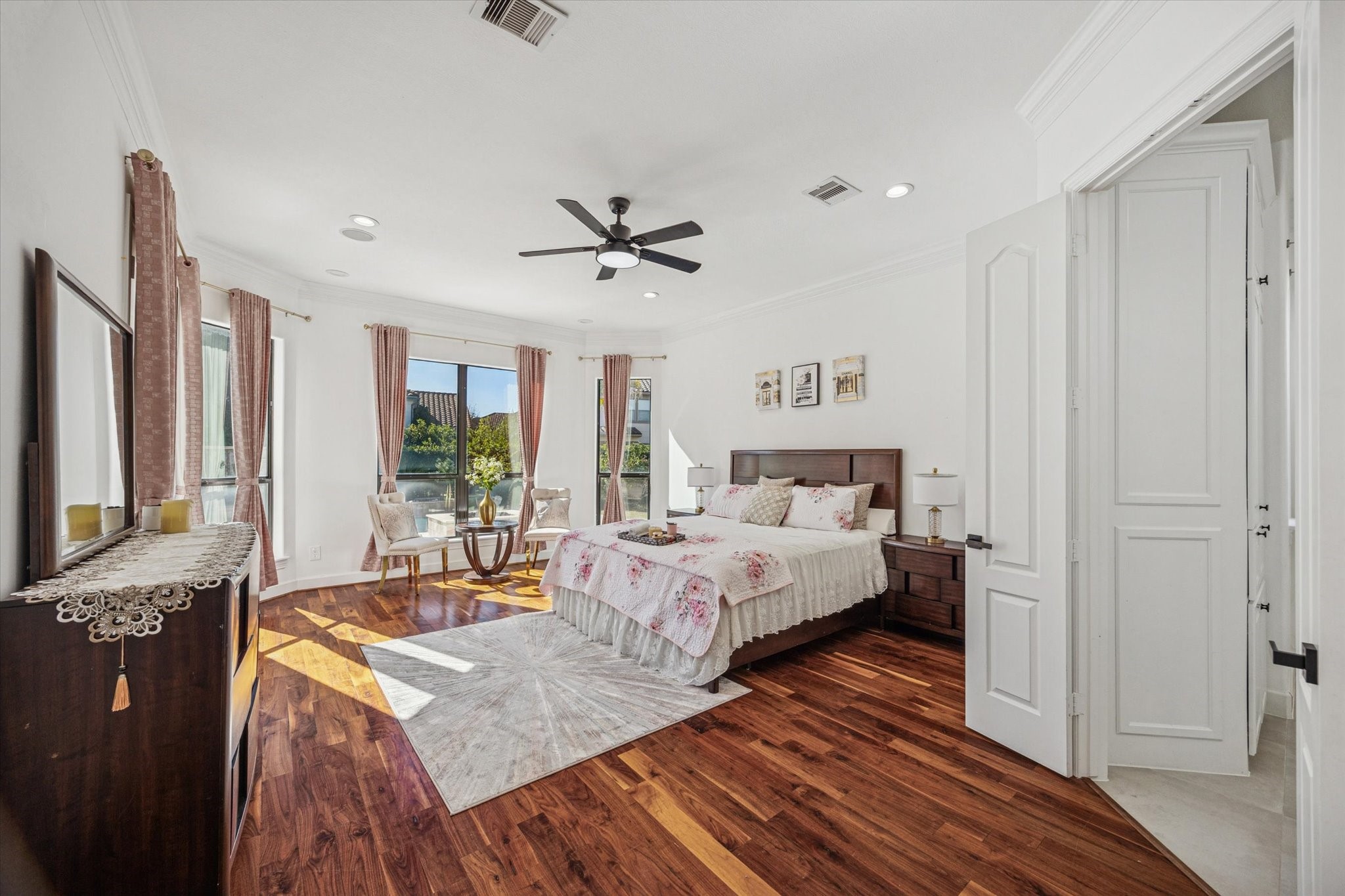 This spacious, bright primary bedroom features hardwood floors, large windows with elegant curtains, and a ceiling fan. It includes a cozy sitting offering a warm and inviting atmosphere.