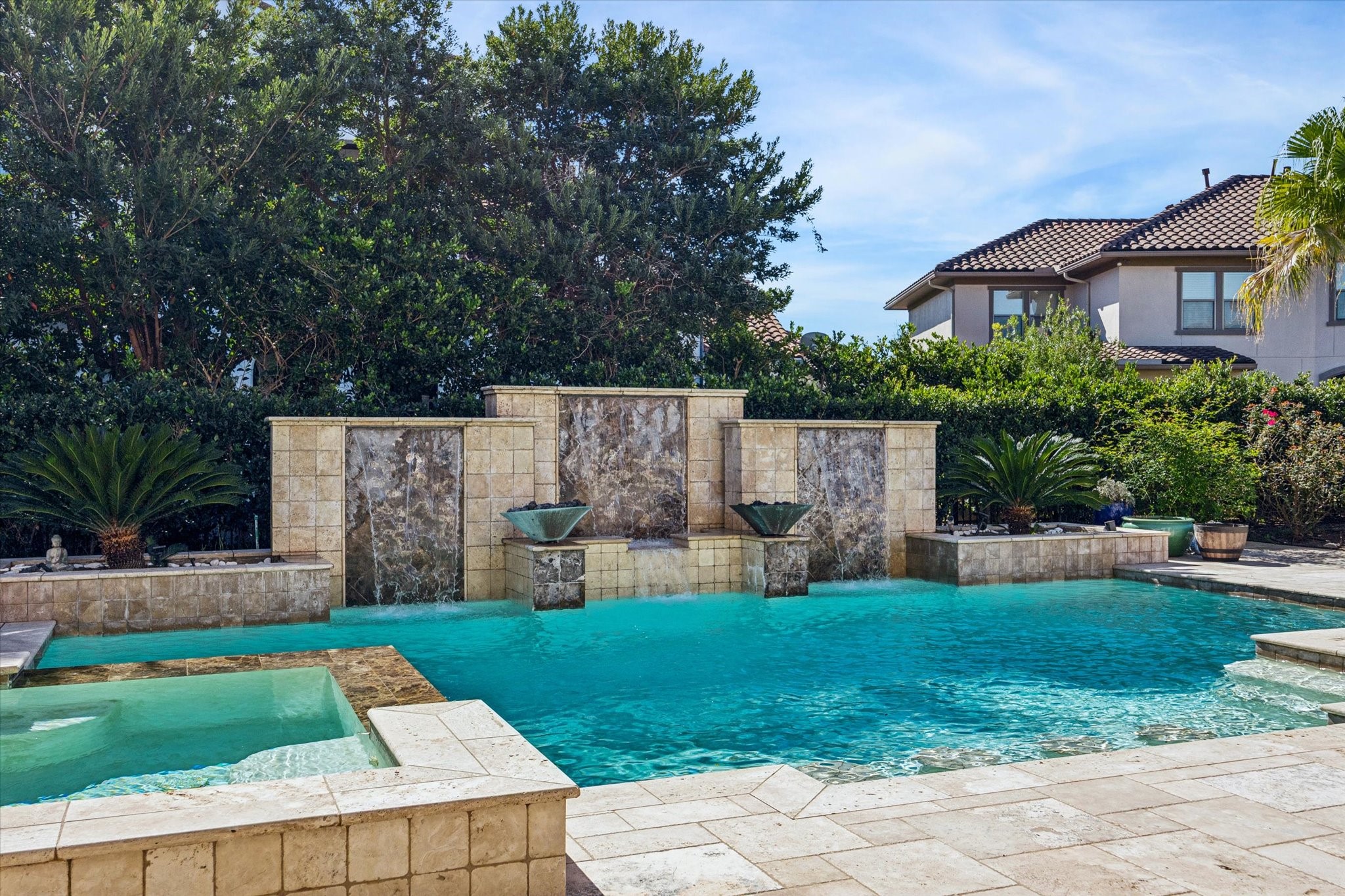 This photo showcases a luxurious backyard with a stunning pool featuring elegant water features and a spa, surrounded by lush greenery and a stylish tile deck, perfect for relaxation and entertainment.