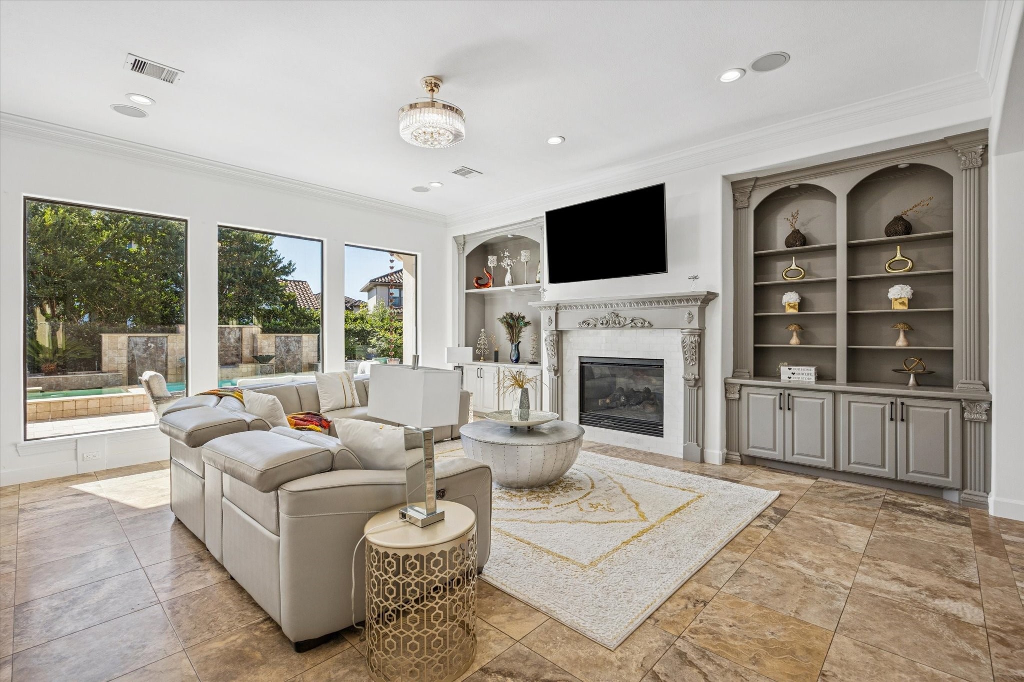 This spacious living room features elegant travertine tile flooring, a cozy fireplace, and built-in shelving. Large windows offer abundant natural light and a view of the outdoor pool area.