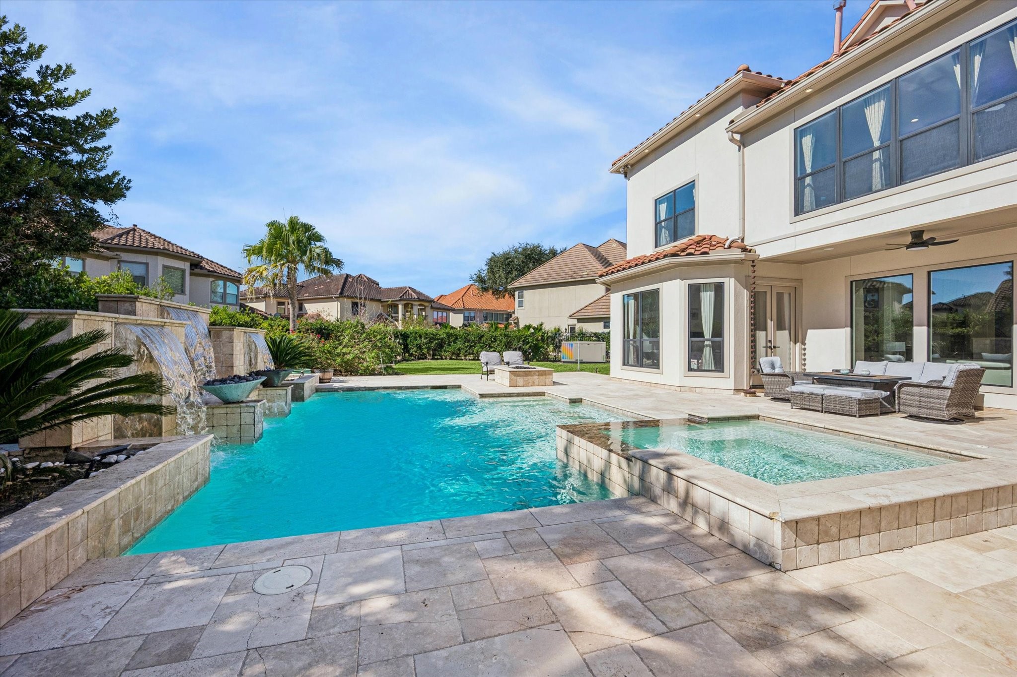This photo showcases a luxurious outdoor space with a sparkling pool and spa, surrounded by elegant stone tiles. The modern two-story home features large windows and a spacious patio area with comfortable seating, perfect for relaxing or entertaining.