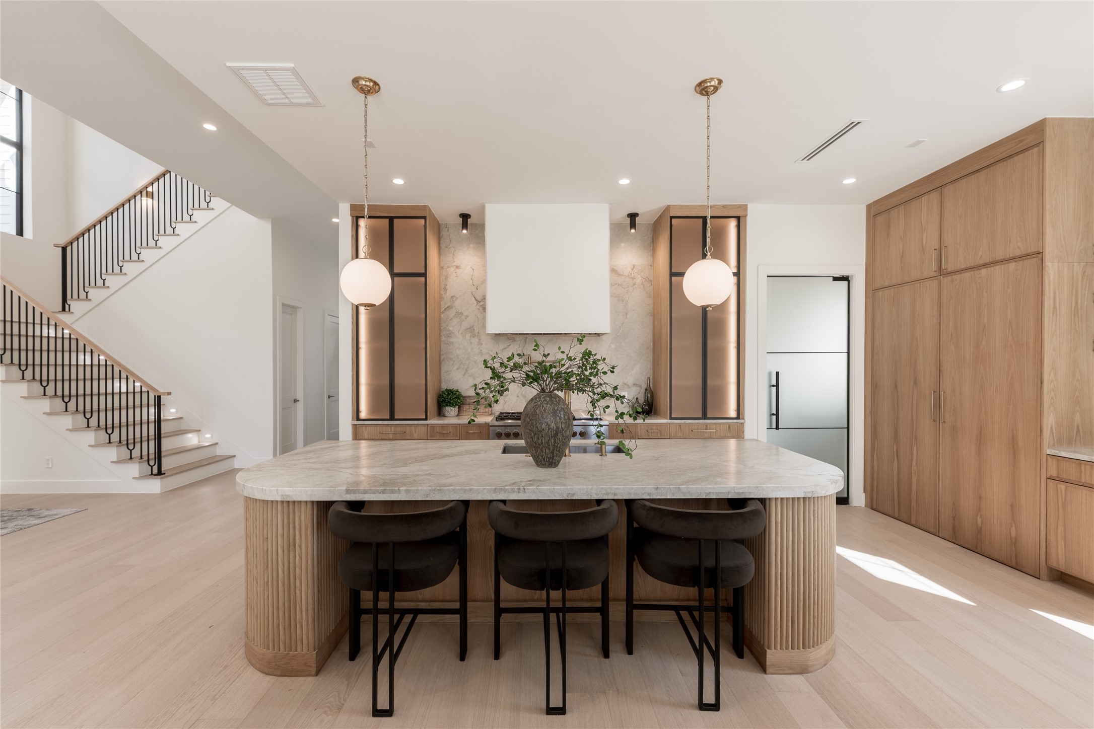 Thoughtfully layered materials create a kitchen that feels both inviting and architecturally distinctive. The expansive stone-topped island anchors the space, complemented by custom white oak cabinetry, softly illuminated displays, and elegant pendant lighting that balances warmth with contemporary design.