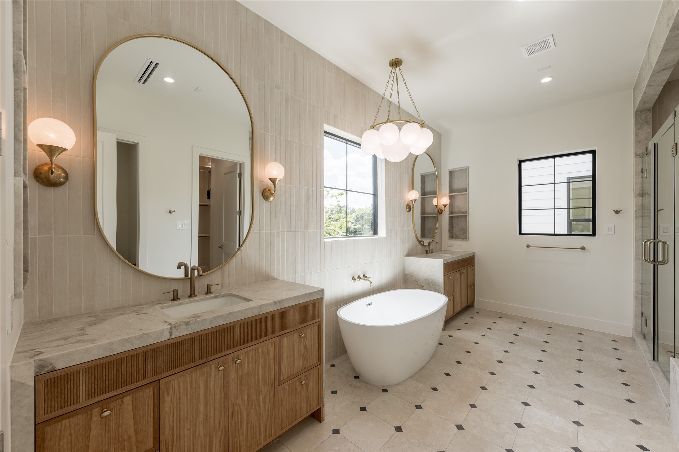 A wide perspective of the primary bath reveals thoughtful symmetry, pairing dual vanities with a freestanding soaking tub and natural stone surfaces. Designer tile carries across the floor and walls, giving the ensuite a tailored, cohesive finish.