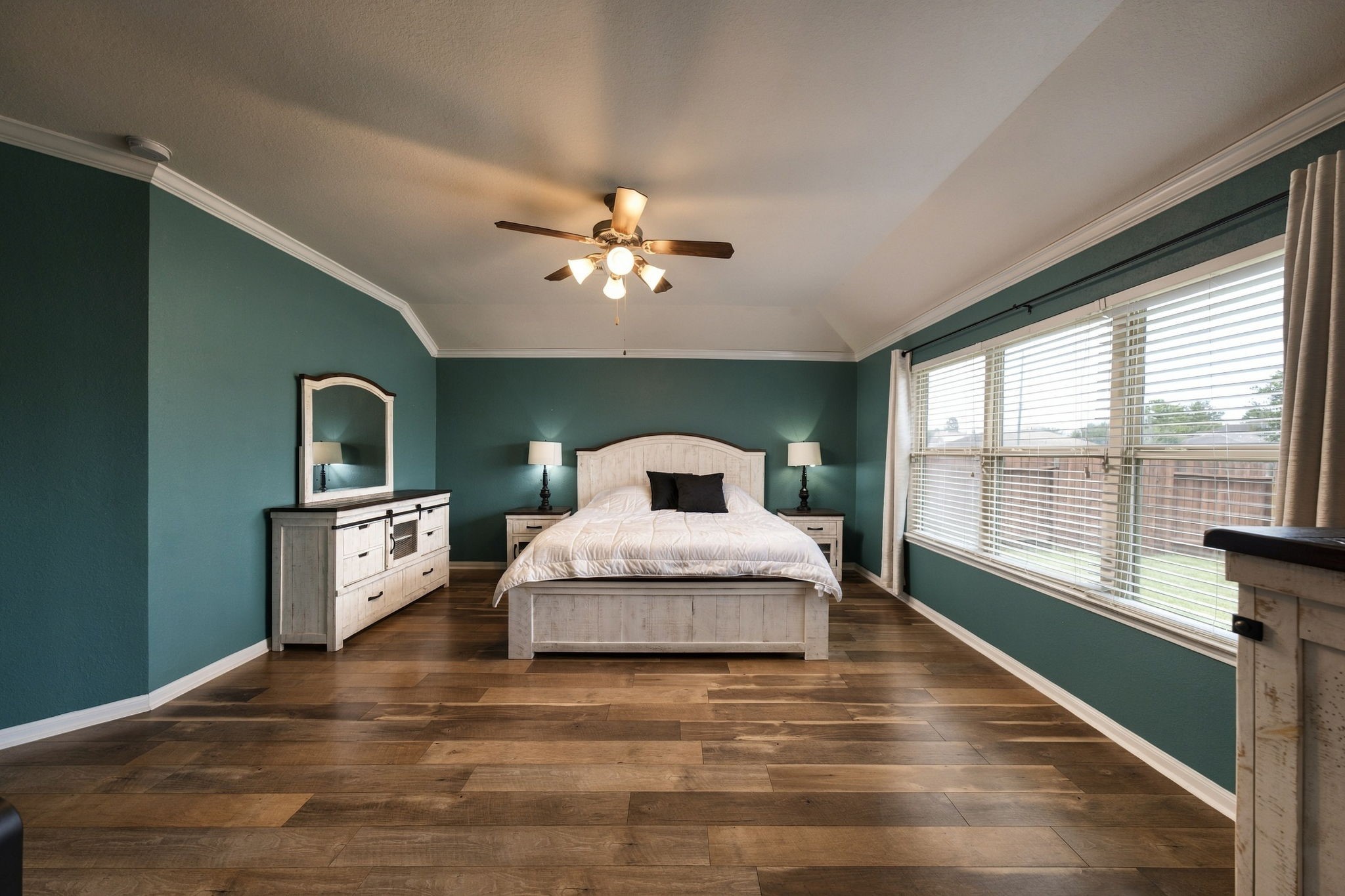 Spacious bedroom featuring rich wooden flooring, a large window for natural light, and elegant teal walls complemented by classic white.