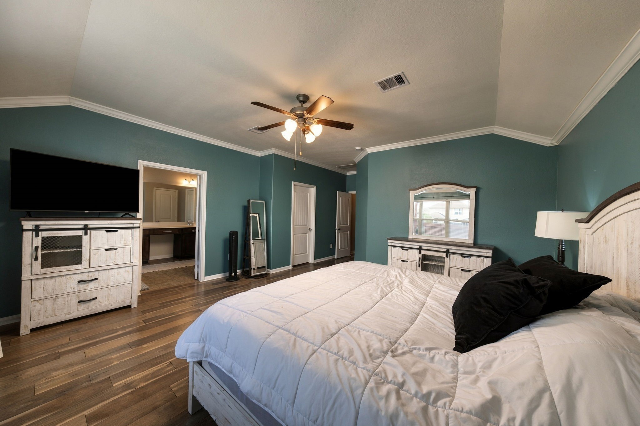 Spacious bedroom with rich teal walls, elegant crown molding, and warm wood flooring. Features include a ceiling fan, ample natural light from a large window offering a cozy and inviting retreat.