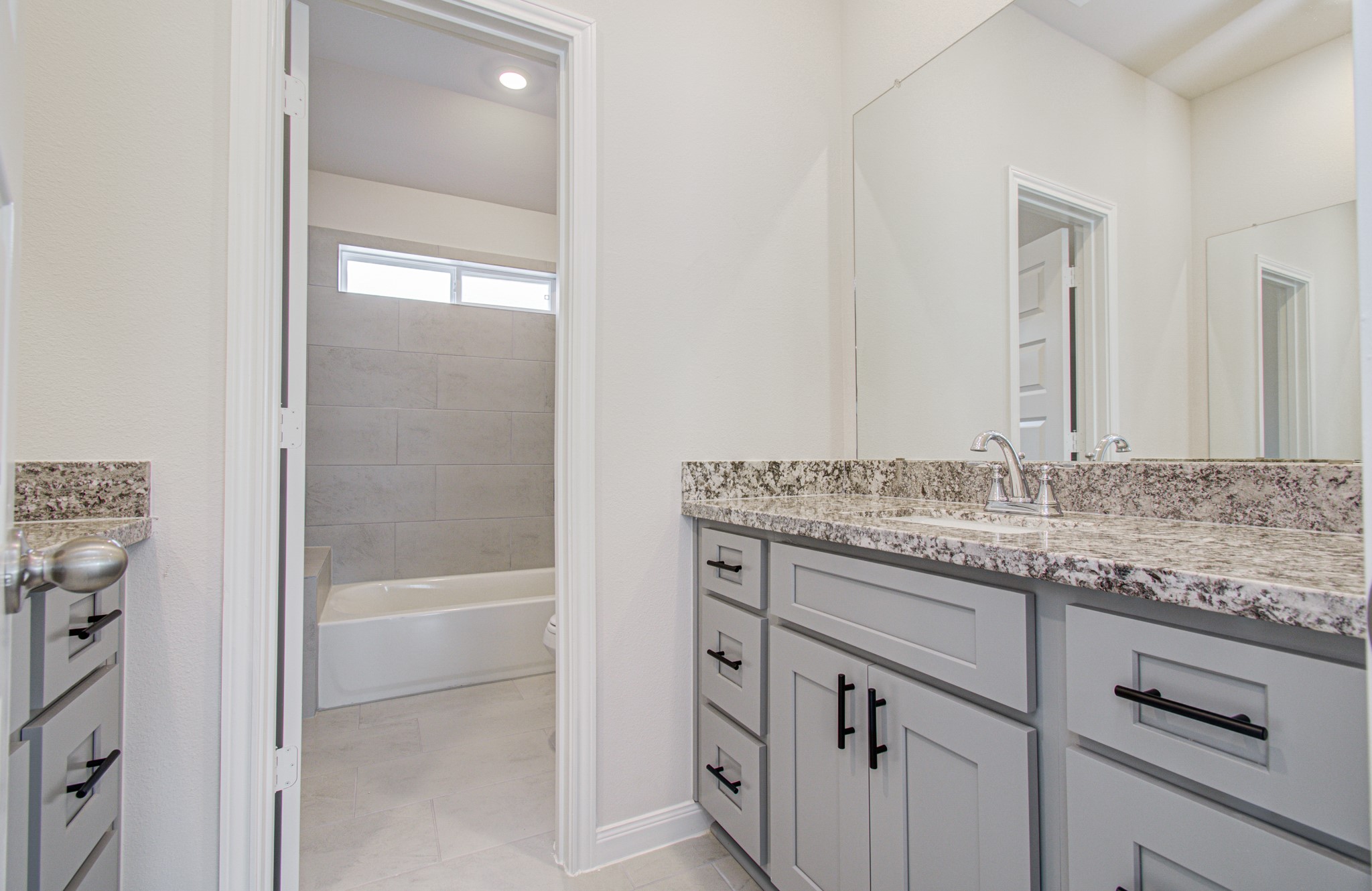 Elegant bathroom featuring a spacious granite-topped vanity with sleek gray cabinetry, leading to a bright tiled tub area with natural light from a high window. Please Note this is a different home/floorplan in the community.