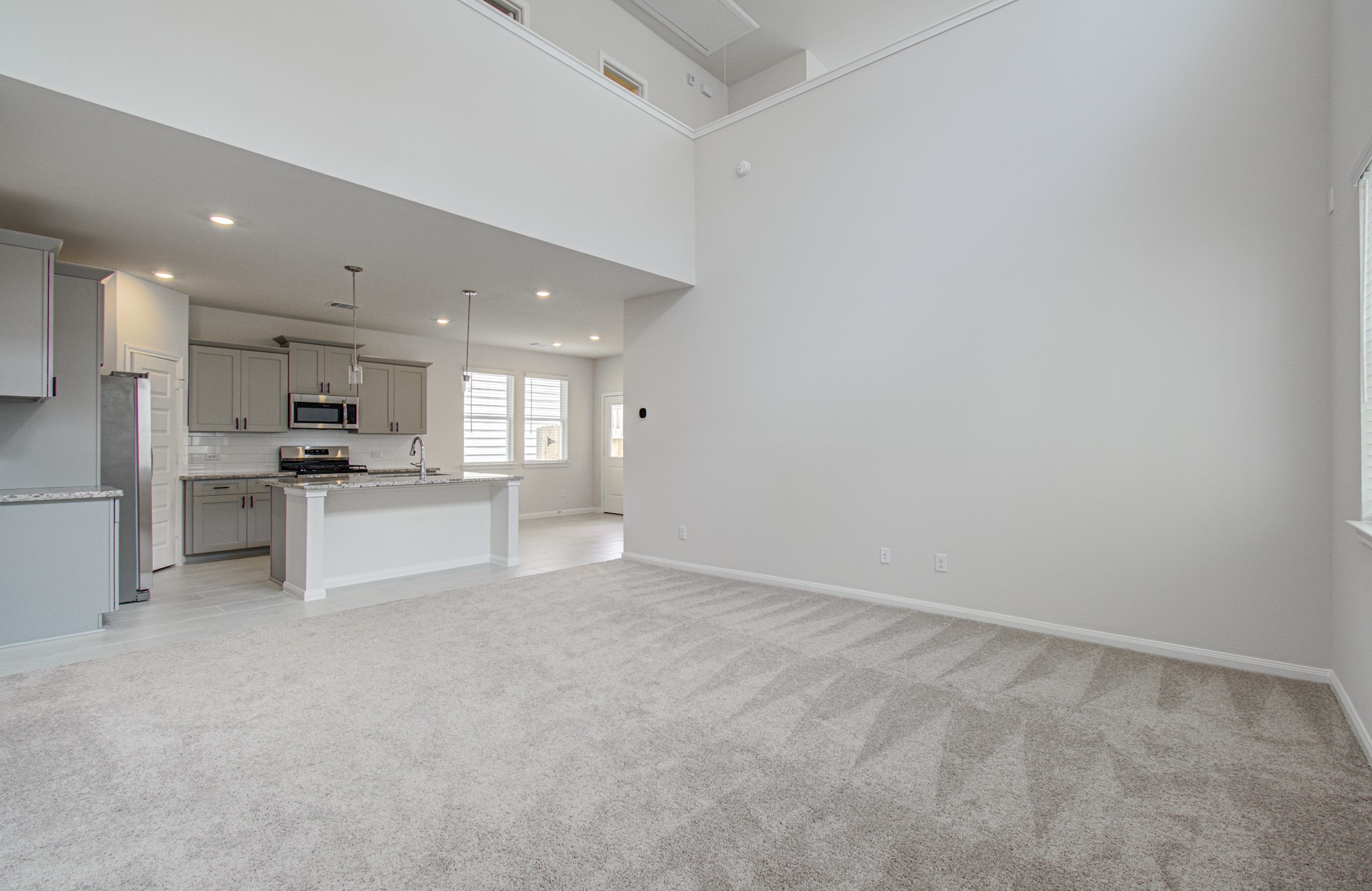 Spacious living area with high ceilings and an open layout, leading to a modern kitchen featuring stainless steel appliances and a large island. Please Note this is a different home/floorplan in the community.