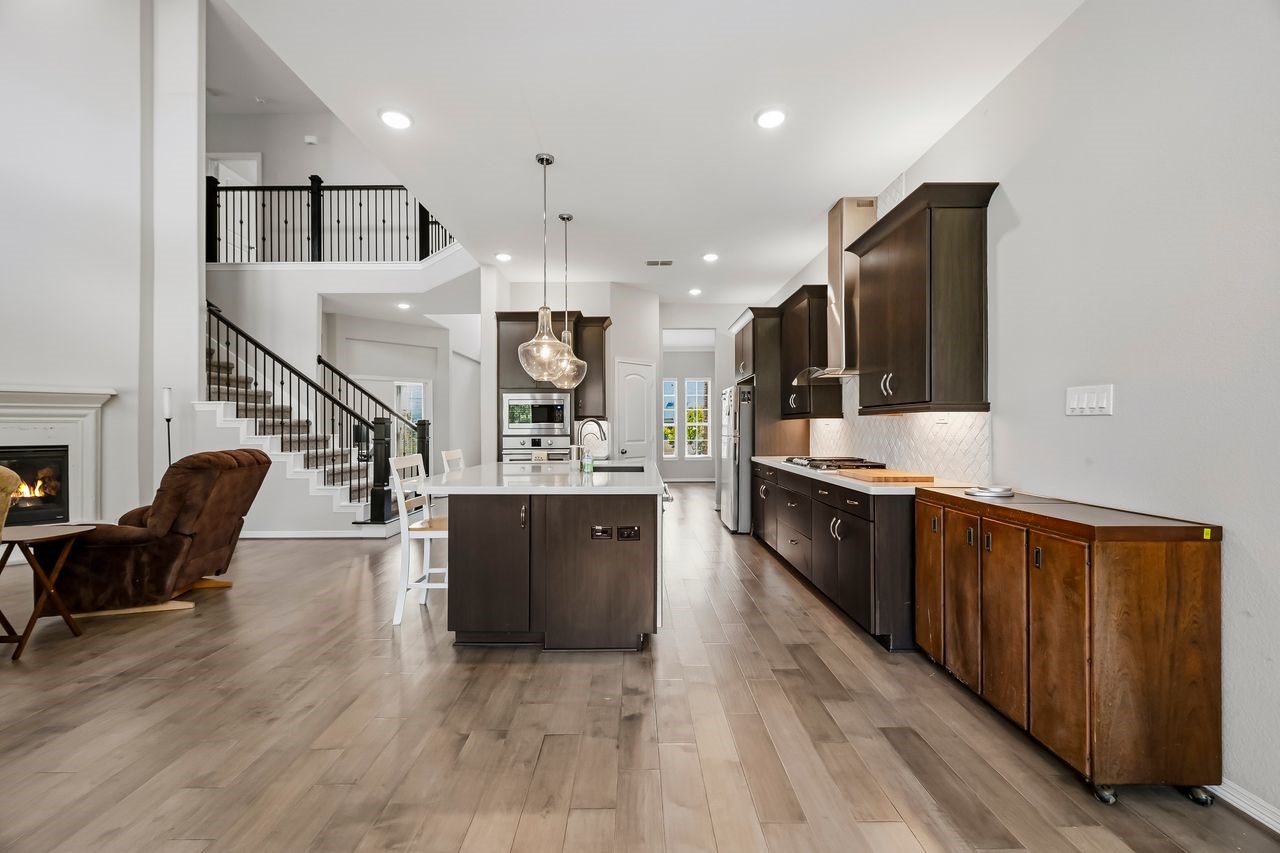 A secondary perspective from the sunlit breakfast bay highlights the seamless flow between the kitchen and living spaces. Dark wood cabinetry and stainless steel appliances provide a sophisticated contrast to the light flooring, while a generous island anchored by elegant pendant lighting serves as the heart of the home. Modern, warm, and perfectly balanced.