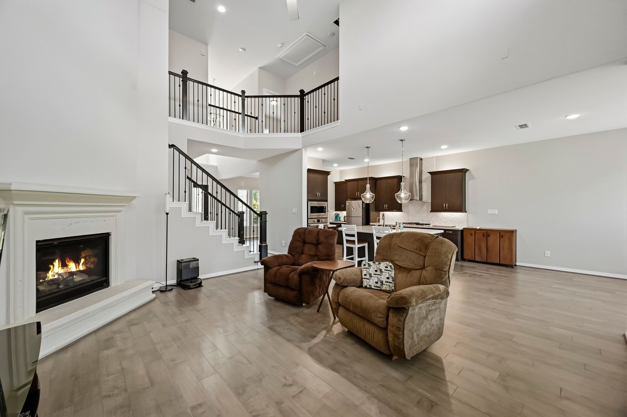 This sophisticated open-concept space seamlessly integrates the living area, breakfast nook, and kitchen, anchored by a contemporary fireplace and a stylish staircase leading to the upper catwalk. Neutral finishes and bold dark accents complement the rich wood flooring, creating a cohesive and inviting vista from the primary retreat entrance.