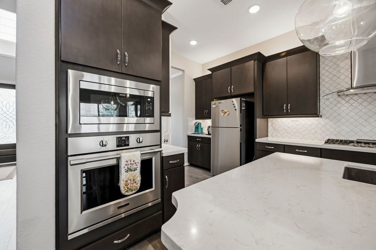 This sophisticated kitchen centers on high-end functionality, showcasing built-in stainless steel ovens set against rich, dark cabinetry. A sleek professional range hood anchors the space, while expansive light-toned countertops with delicate grey veining offer ample room for preparation. Tucked beyond the main workspace, a dedicated breakfast and coffee nook completes this refined culinary environment.