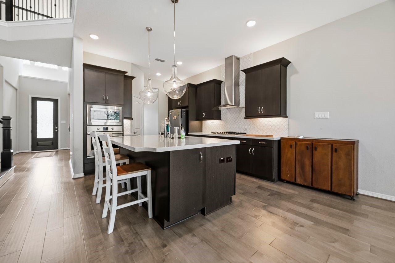 Looking through to the entryway, this contemporary kitchen makes a sophisticated statement with its rich, dark cabinetry and sleek tile backsplash. The space is thoughtfully designed with under-cabinet lighting and a suite of high-end stainless steel appliances, including a gas cooktop and built-in microwave.