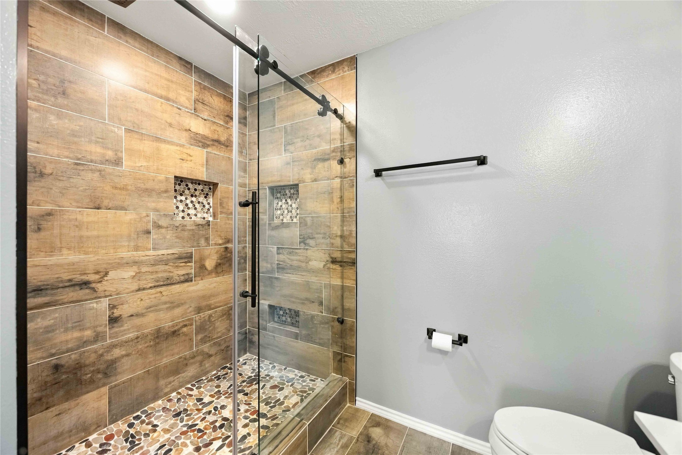 Custom walk-in shower is finished with wood-look tile, pebble-stone flooring, built-in niches, and sliding glass enclosure.