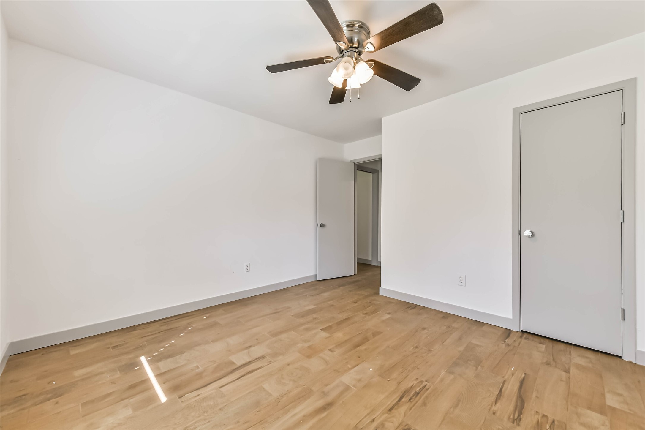 Bedroom features light wood flooring, white walls, and a ceiling fan with lights. It has two doors and ample natural light, creating a bright and airy space.