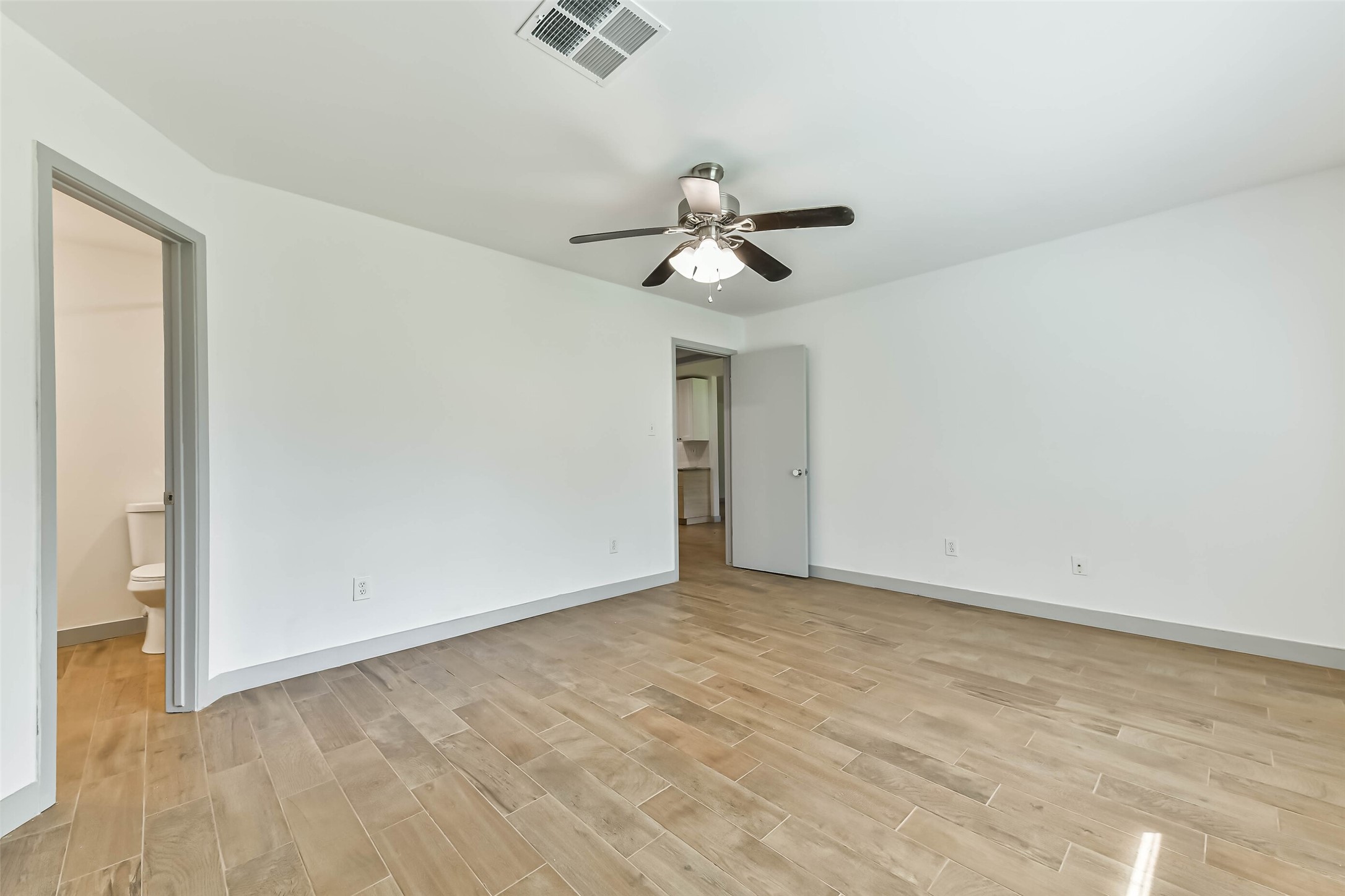 Bright, spacious bedroom with light wood flooring, a ceiling fan, and a neutral color palette. It includes an en-suite bathroom and a closet, offering a comfortable and functional living space.