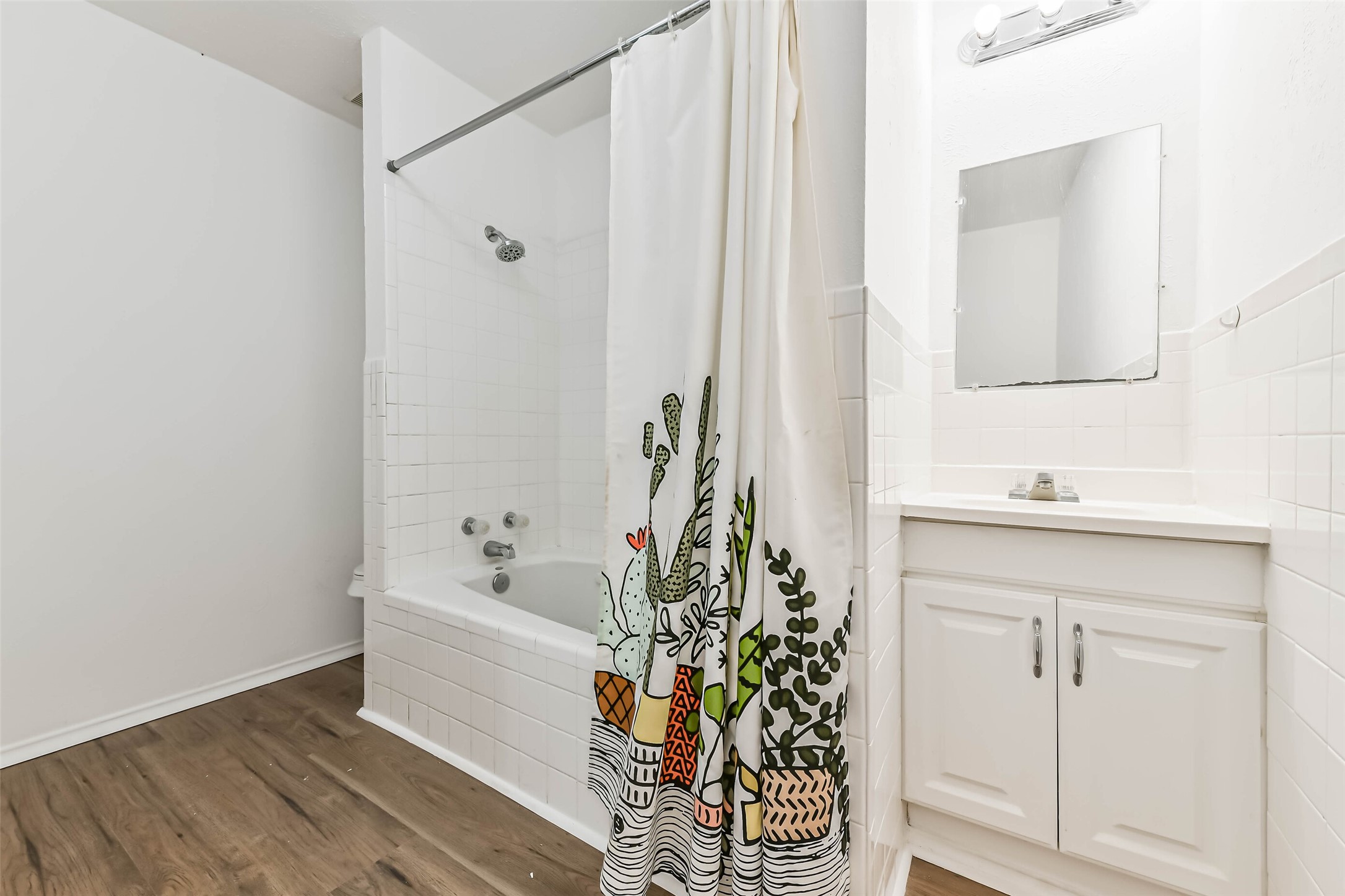 Bathroom features a clean, white design with a tiled shower and tub. It includes a vanity with storage, a large mirror, and wood-style flooring for a warm touch.