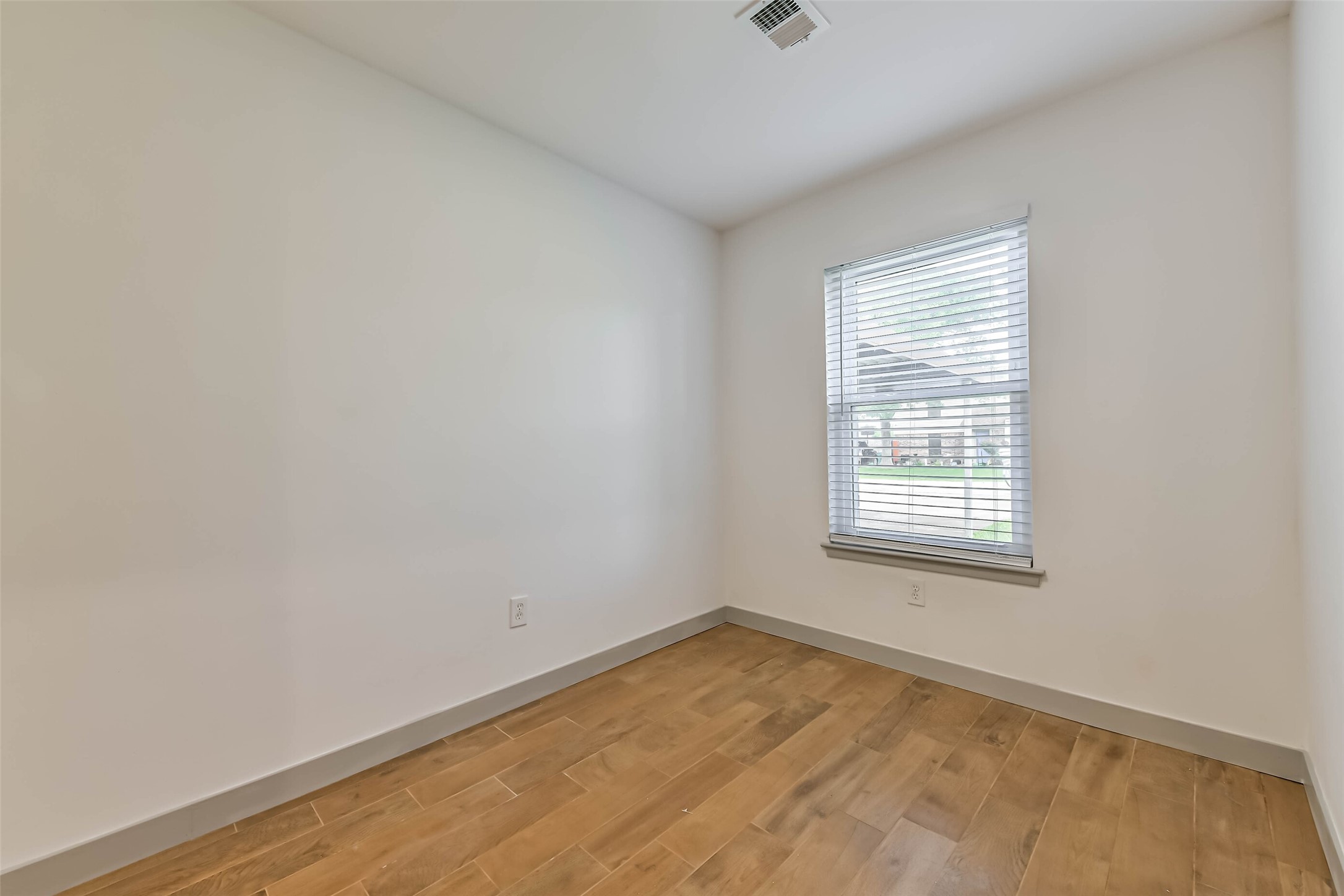 A bright room with white walls and light wood flooring, featuring a window with blinds that lets in natural light. Perfect for a cozy office or guest room.