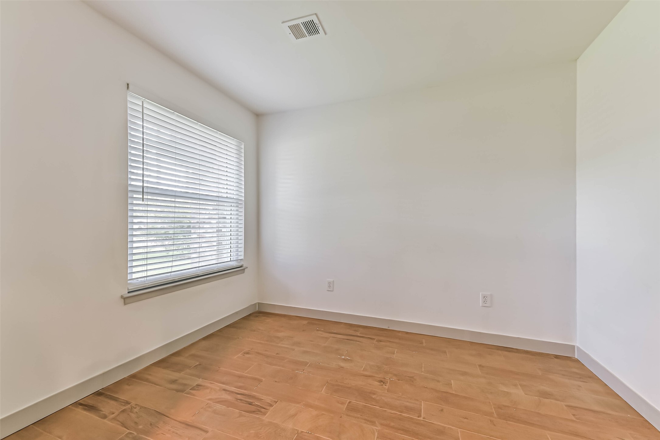 Bedroom with clean white walls and a large window with blinds. The space features light wood flooring and a ceiling vent, offering a blank canvas for customization.