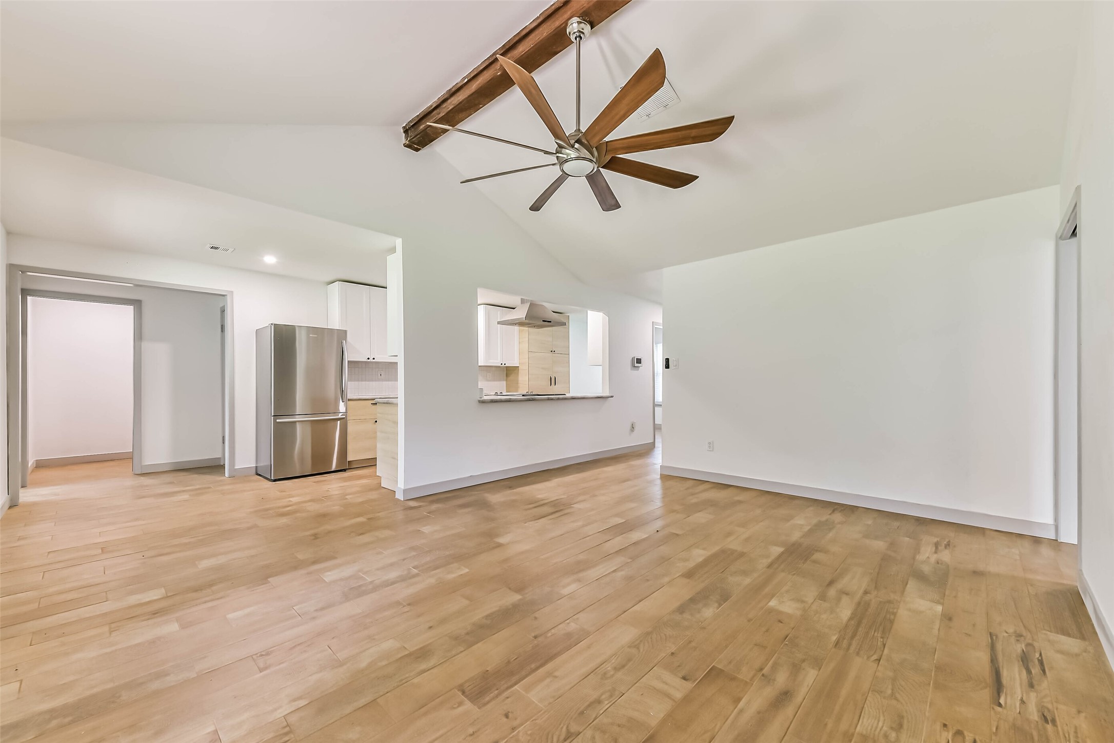 Open living space with light wood flooring and a ceiling fan. The room flows into a kitchen area with stainless steel appliances, including a refrigerator, and a partial view of white cabinetry.
