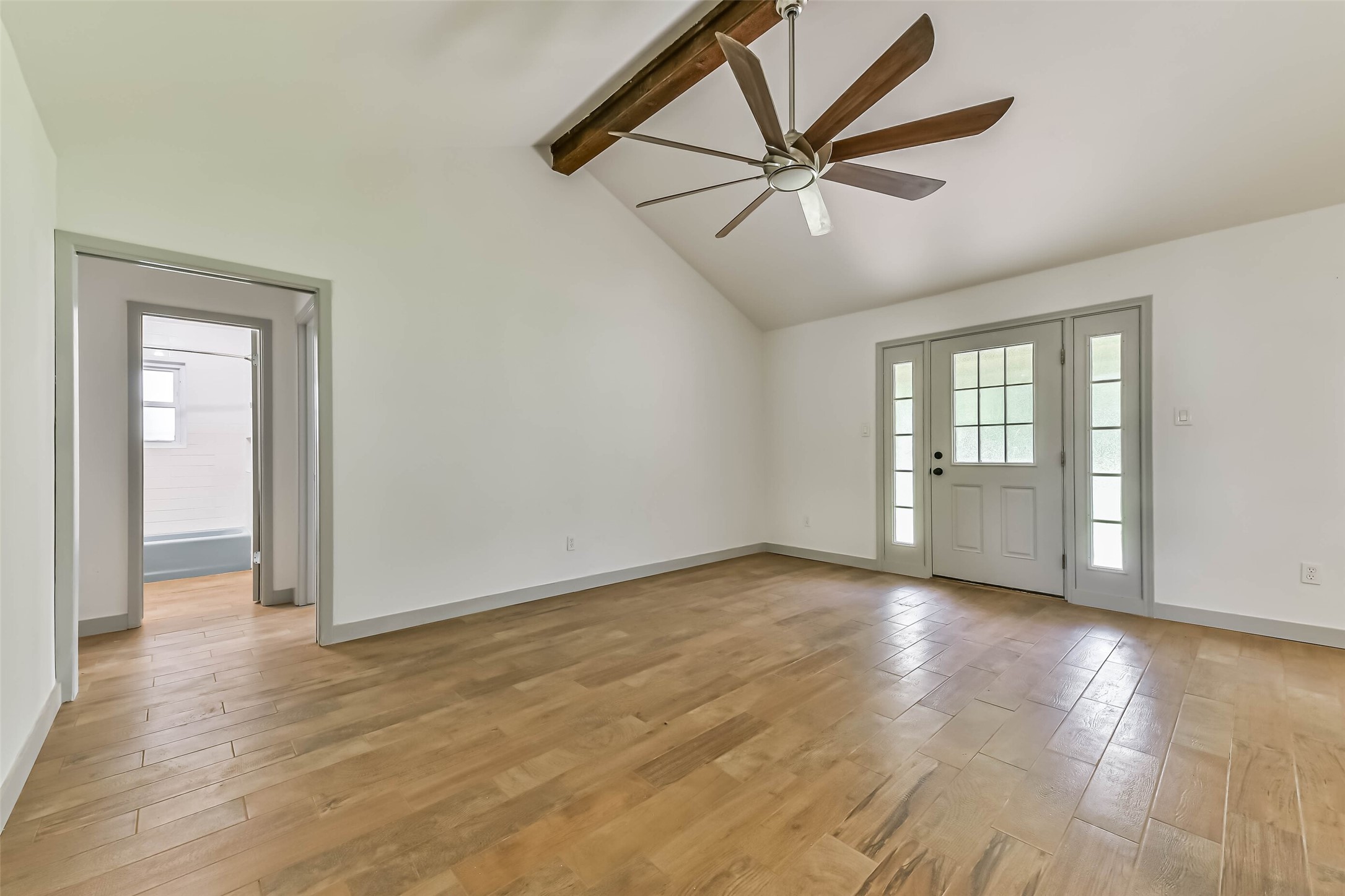Vaulted ceiling with a wooden beam and ceiling fan, hardwood floors, and a neutral color scheme. It has French doors leading outside and an adjoining bathroom, providing a spacious, airy feel.