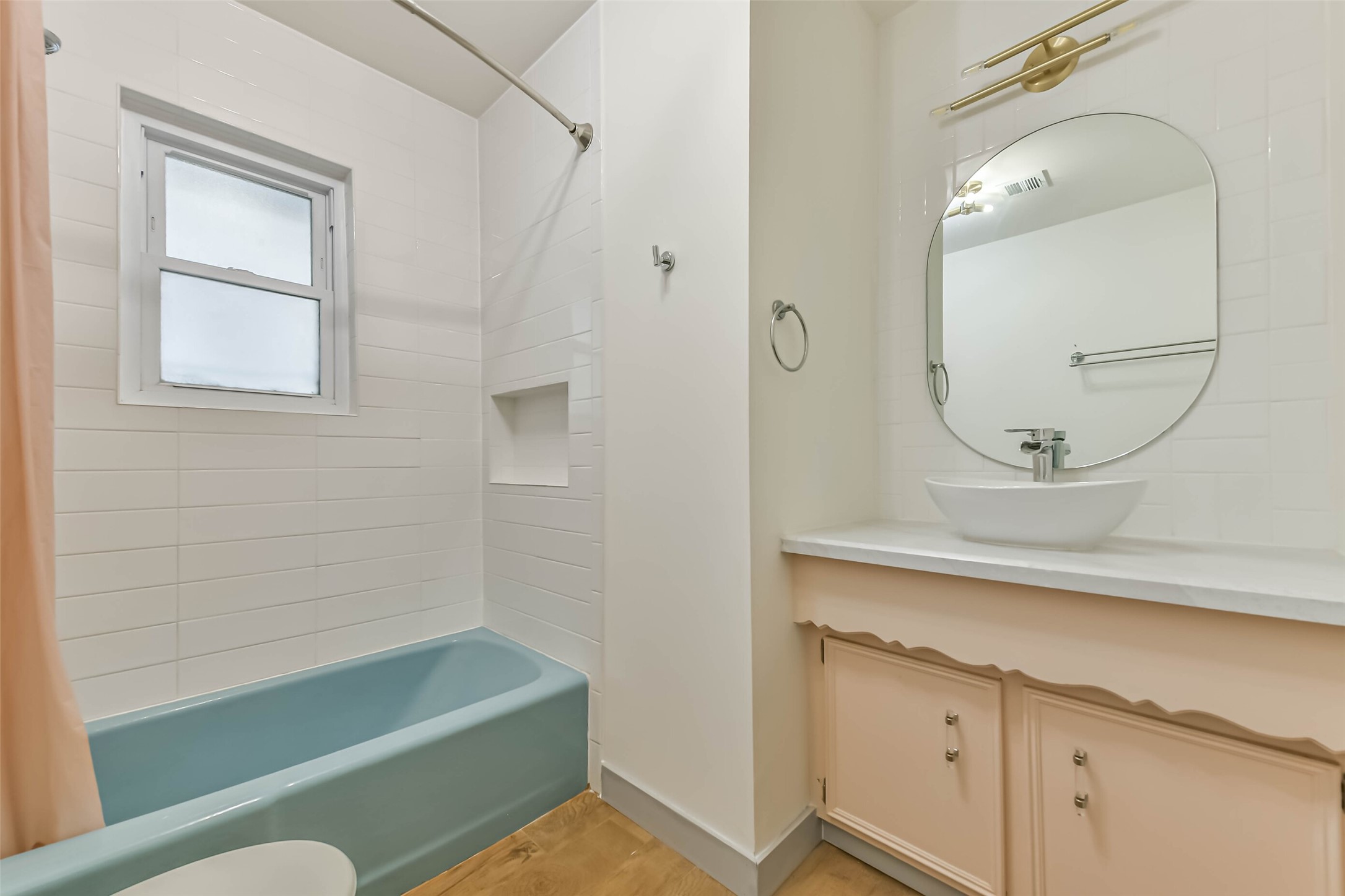 Bathroom features a soothing blue bathtub with white tile surround, a modern vessel sink on a peach vanity, and an oval mirror. A window provides natural light, enhancing the clean, bright atmosphere.