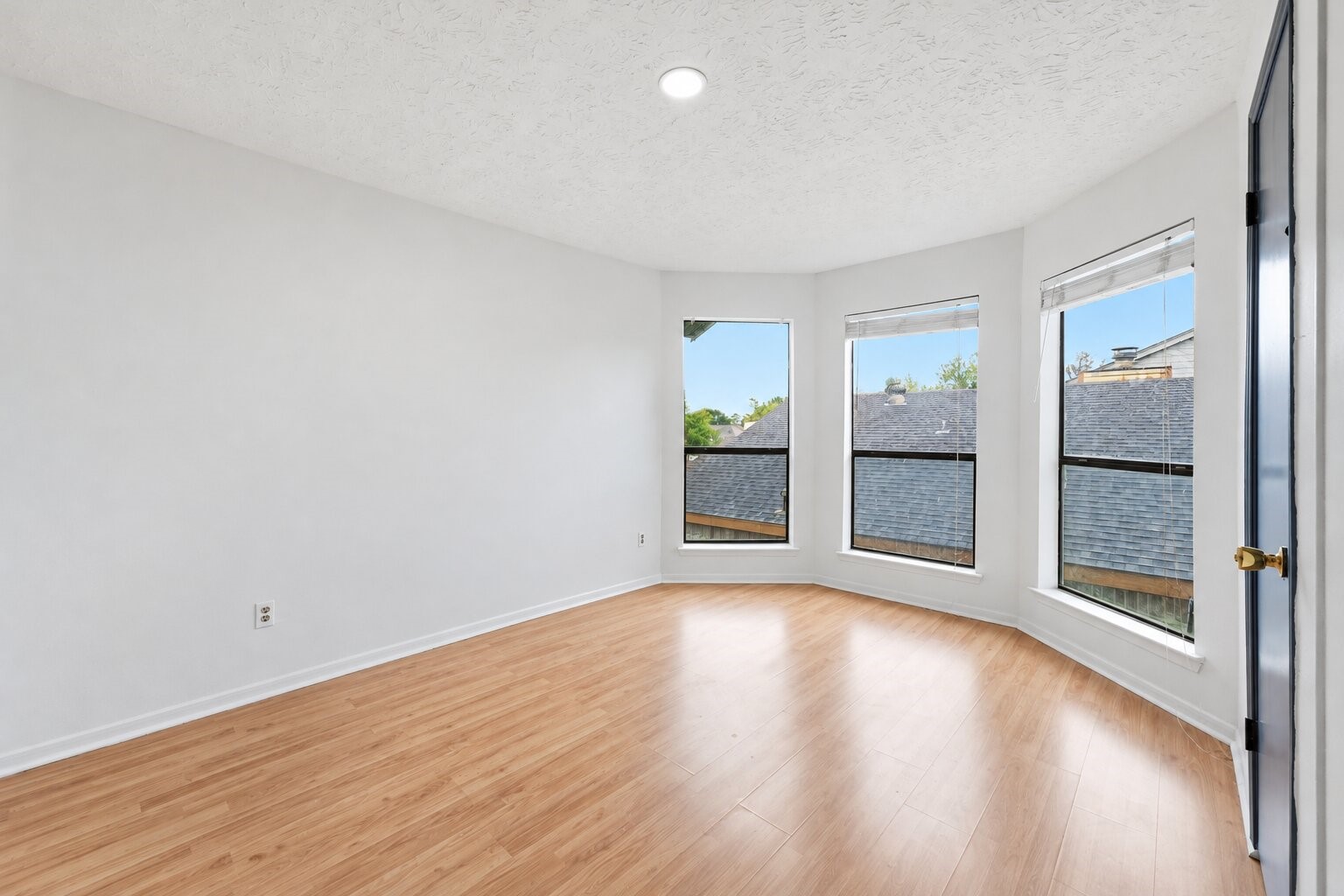 Secondary bedroom welcomes daylight through three angled windows, complemented by wood-look flooring and a flexible layout