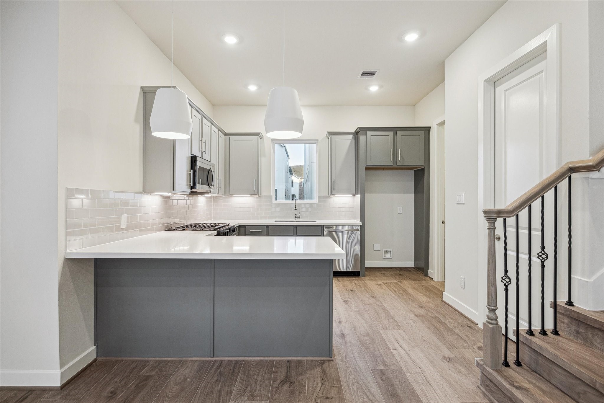 Kitchen featuring a large peninsula, gray cabinetry, white countertops, subway tile backsplash, pendant lighting, stainless steel appliances, and an open connection to the main living area. Kitchen featuring a large peninsula, gray cabinetry, white countertops, subway tile backsplash, pendant lighting, stainless steel appliances, and an open connection to the main living area.