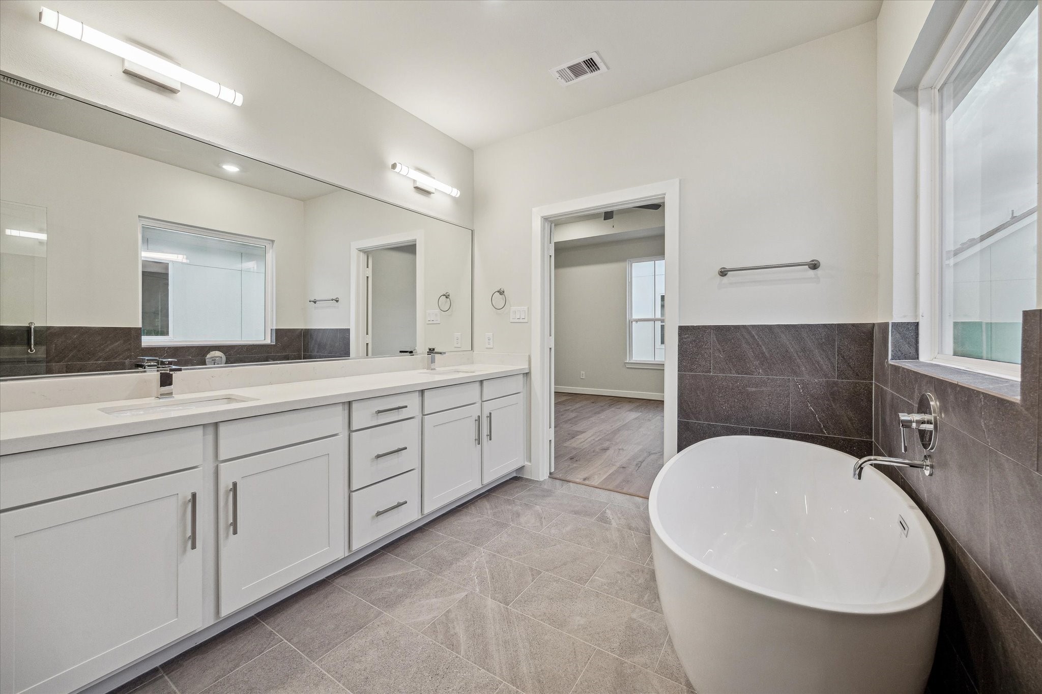 Primary bath featuring a long dual-sink vanity, white cabinetry, large mirror, freestanding soaking tub, modern tile surround, and spacious connection to the primary suite.