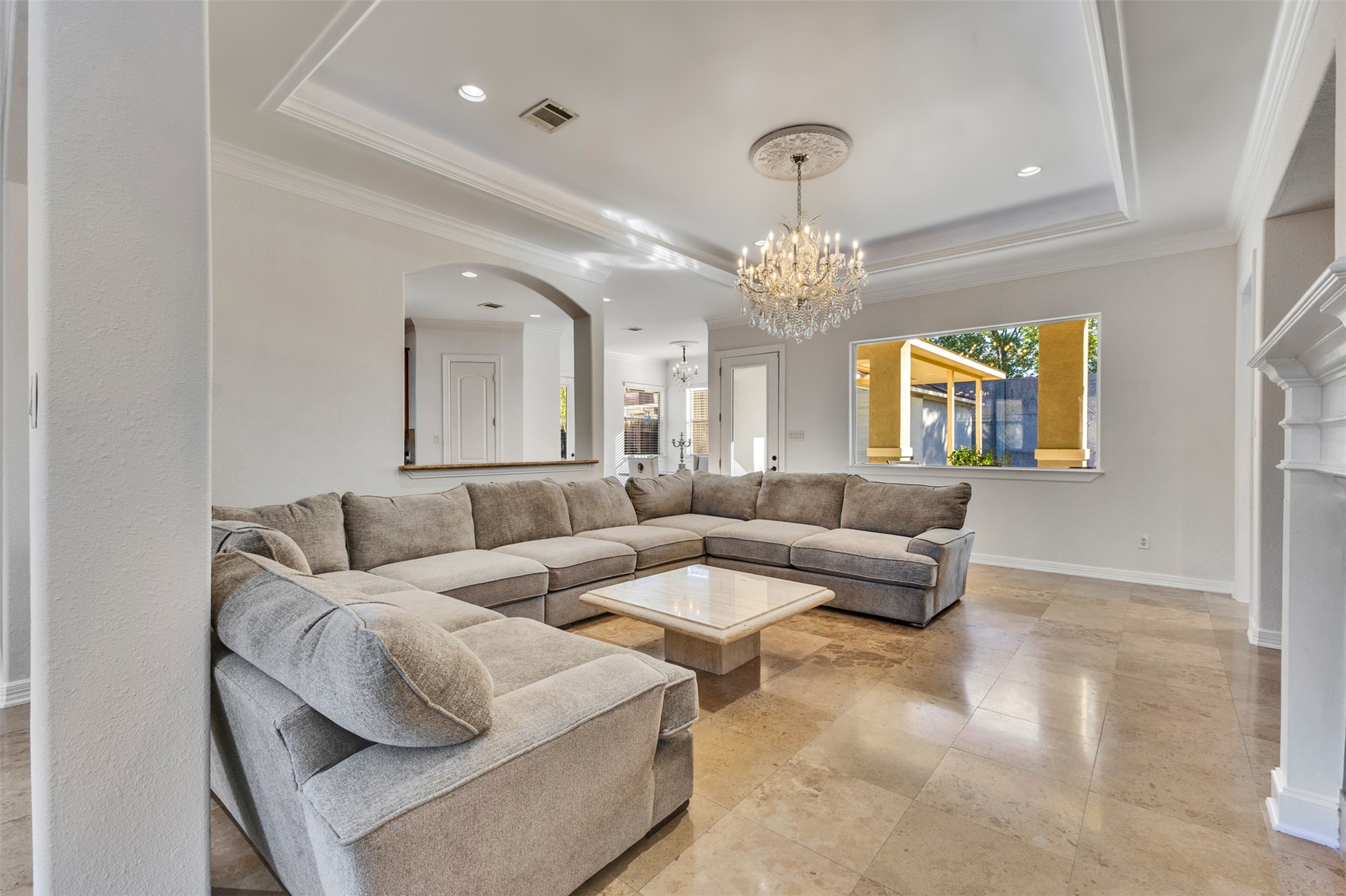 Elegant living room featuring high tray ceilings framed by intricate crown molding, a stunning chandelier with a decorative ceiling medallion, and graceful arched openings. Abundant natural light enhances the spacious, inviting atmosphere.